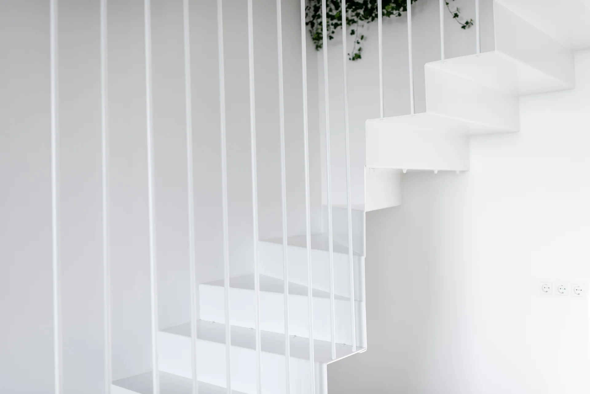 A minimalist white metal staircase features thin vertical rod railings against a plain white wall with green plant foliage visible at the top.