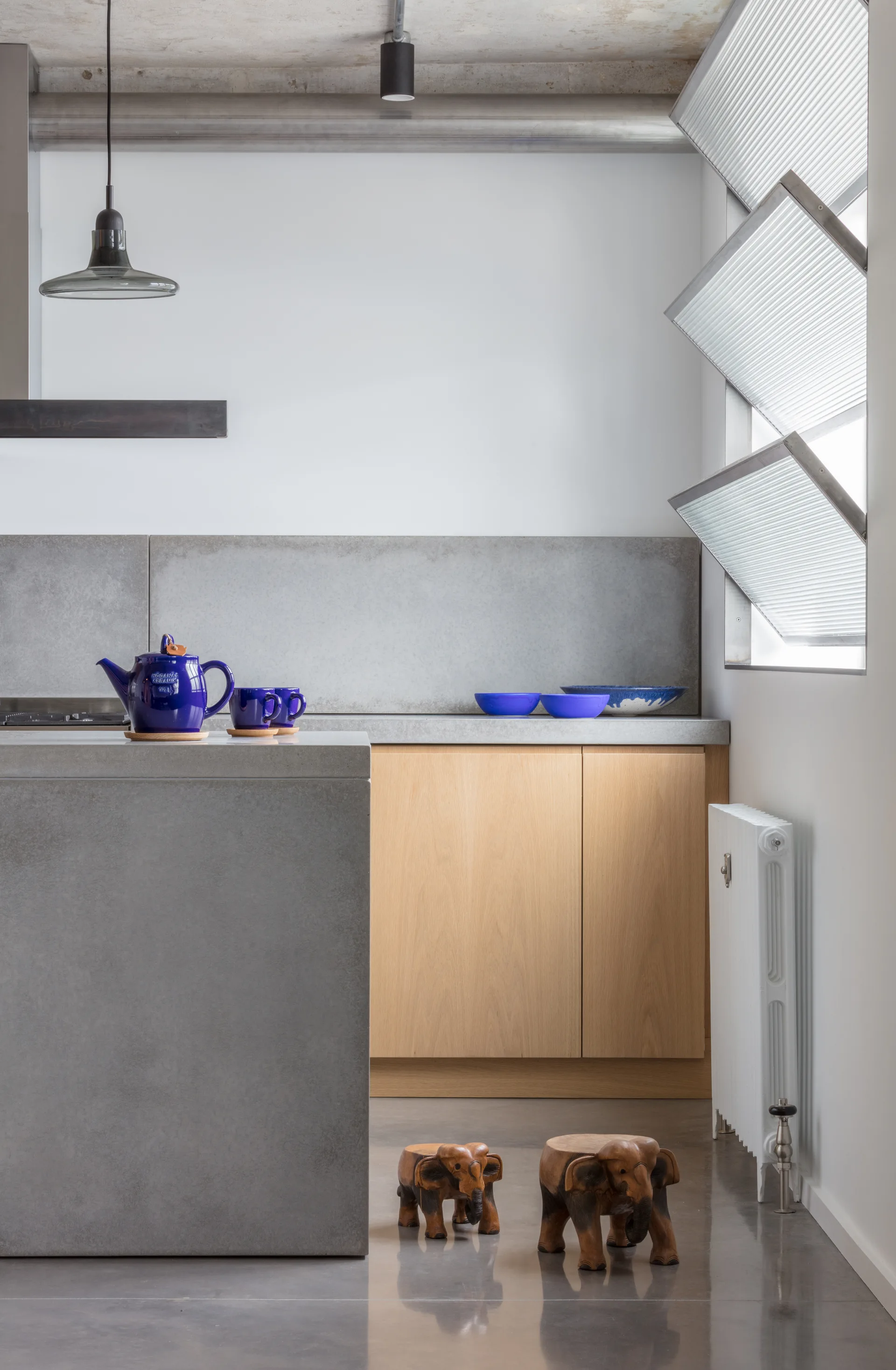 A minimalist kitchen features a grey concrete island, light wood cabinets, and bright blue ceramic dishes on the counter.