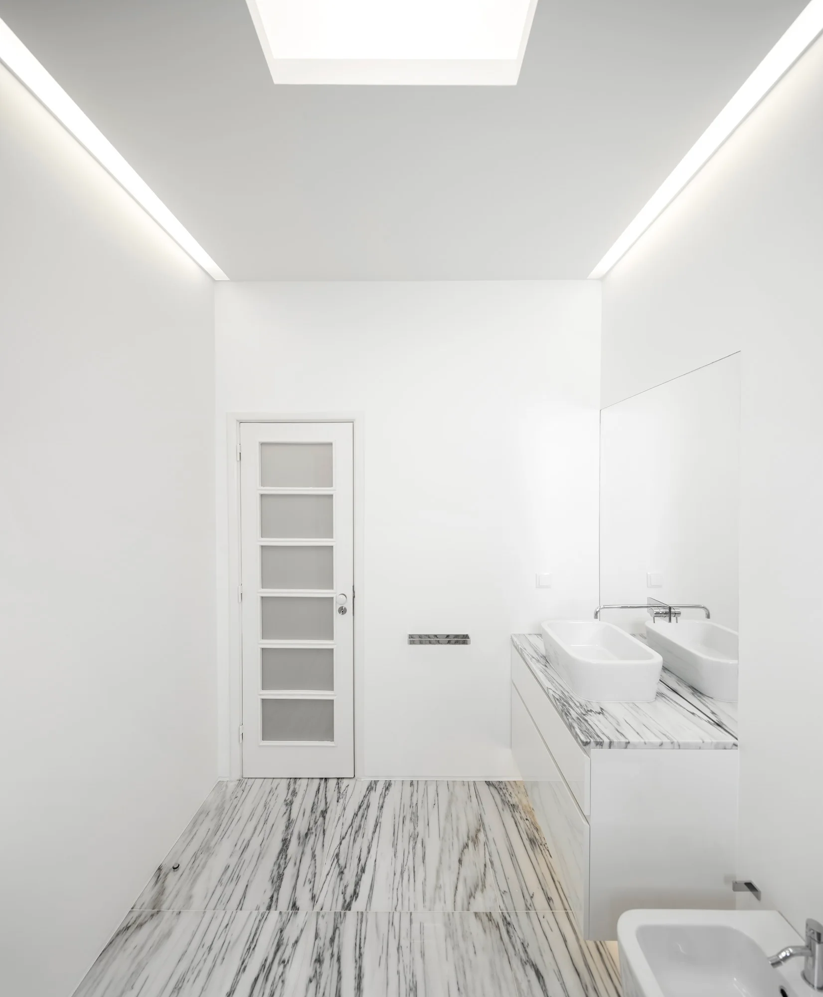 A minimalist white bathroom with heavily veined marble flooring, a floating marble-topped vanity, a vessel sink, and linear ceiling lights.