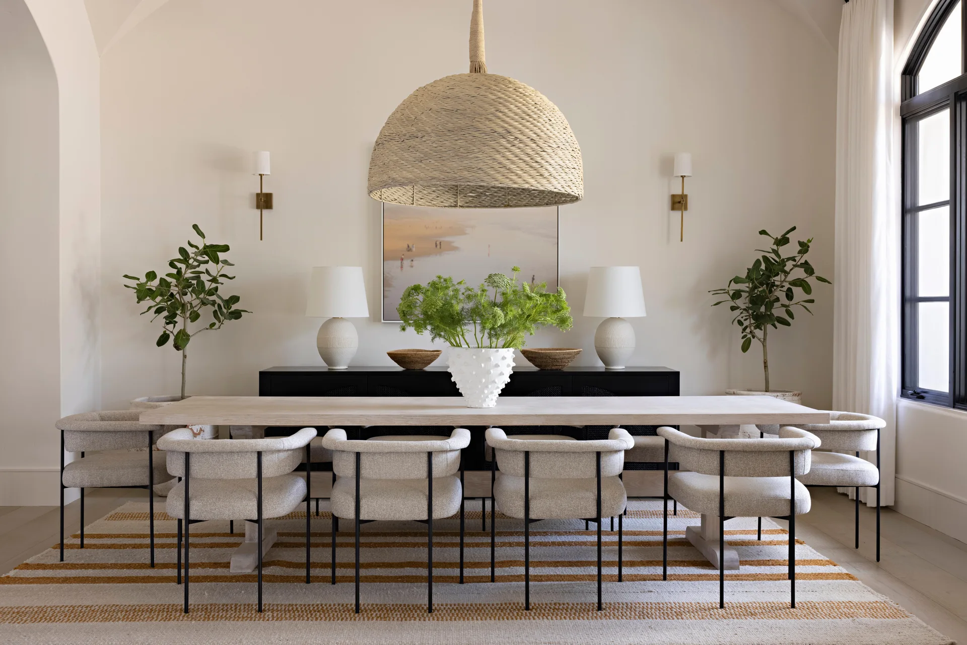 A light wood dining table with cream upholstered chairs sits on a striped rug under a large woven dome pendant light in a bright dining room.