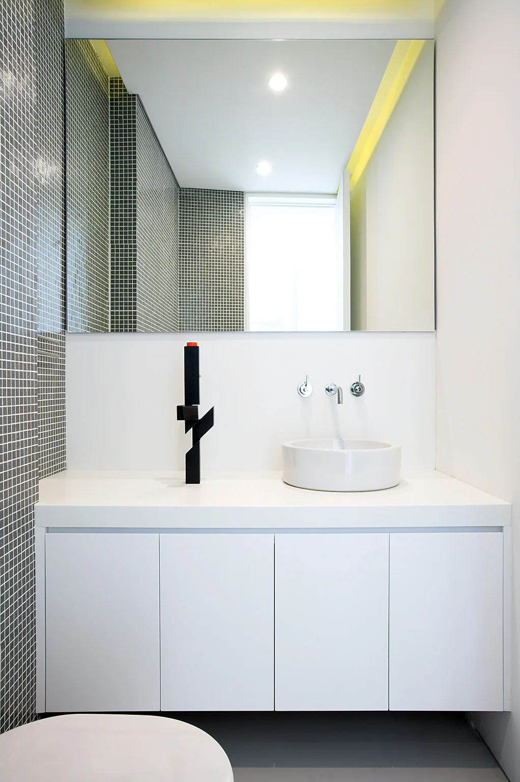 A minimalist bathroom vanity features a round white vessel sink, a wall-mounted faucet, a large mirror, and a black mosaic tile accent wall.