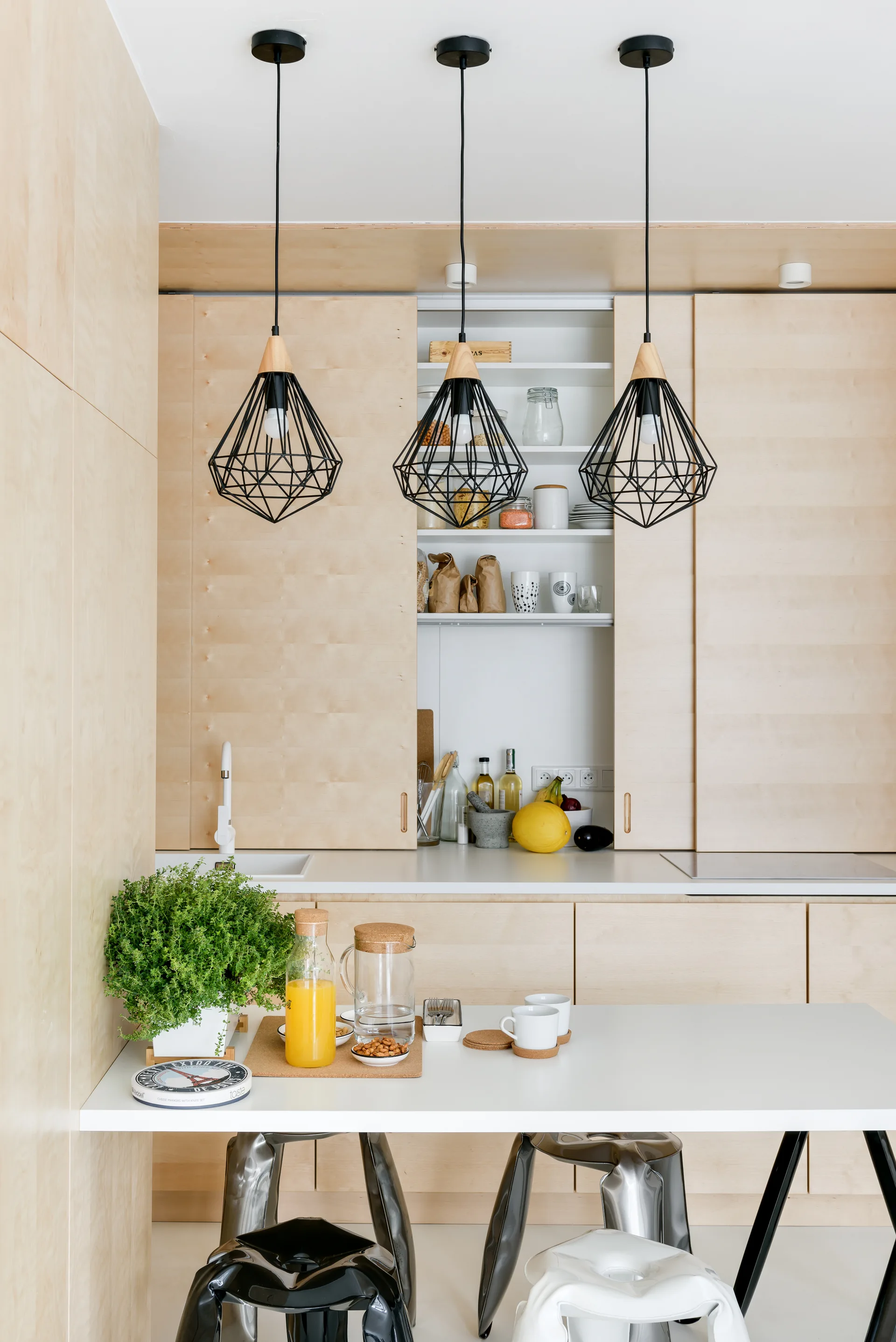 A minimalist kitchen features light plywood cabinets with a sliding pantry door, white countertops, and three black geometric pendant lights.
