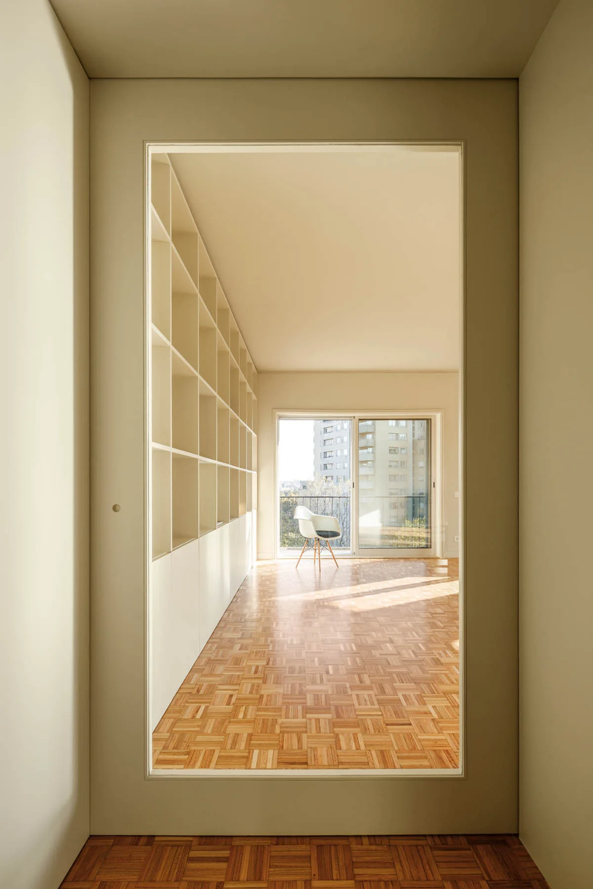 A view through a doorway into a sunlit room featuring parquet wood floors, a full-wall white built-in bookcase, and a single white chair.