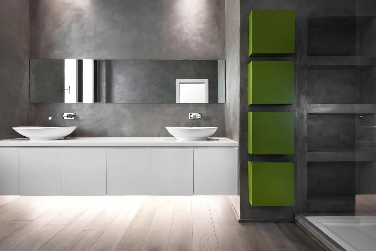 A modern bathroom features a floating white double vanity under a long mirror, textured gray plaster walls, and four bright green square storage cabinets.