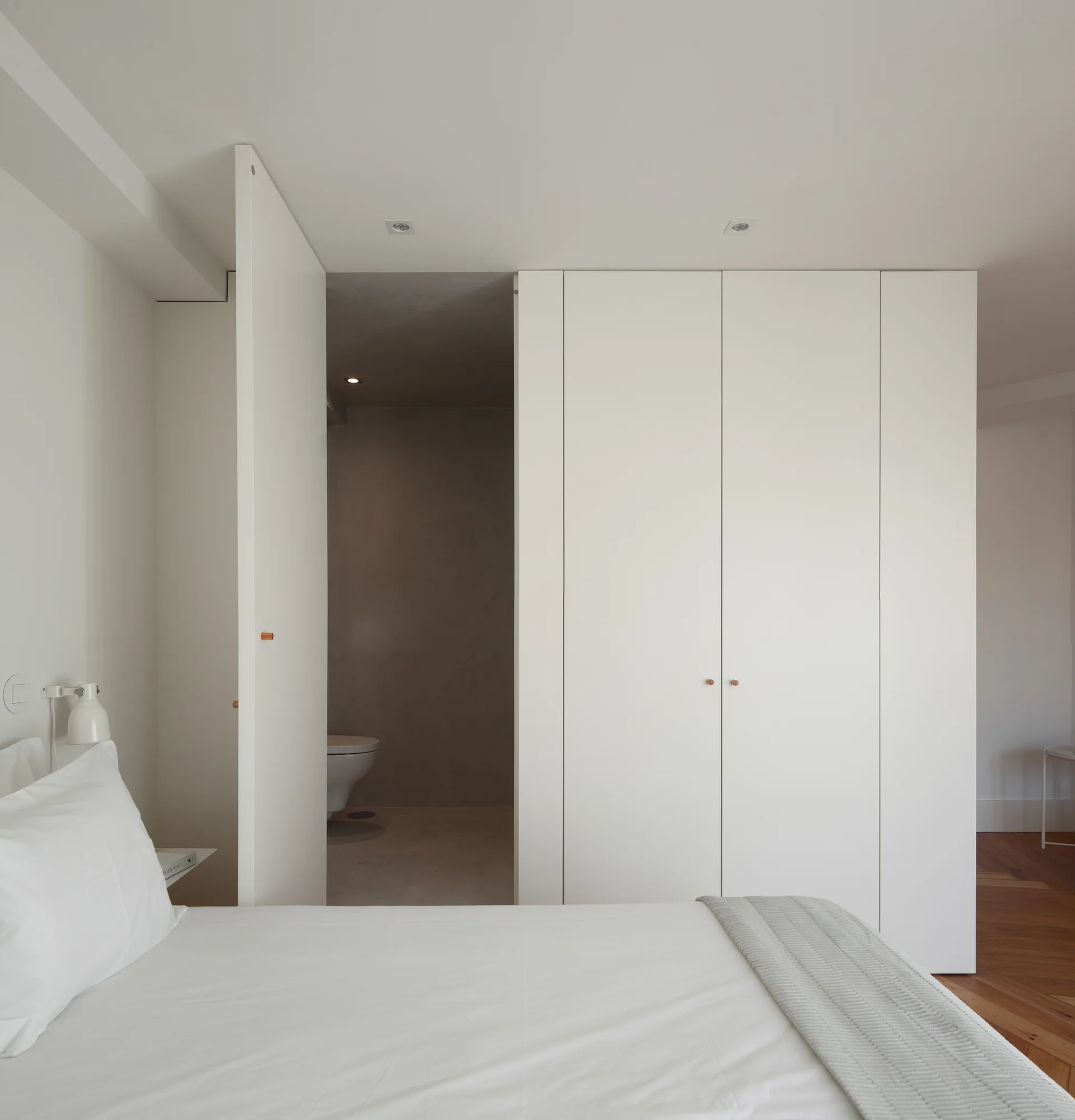 A minimalist white bedroom features a floor-to-ceiling wardrobe with small wooden knobs next to a doorway leading into a grey ensuite bathroom.