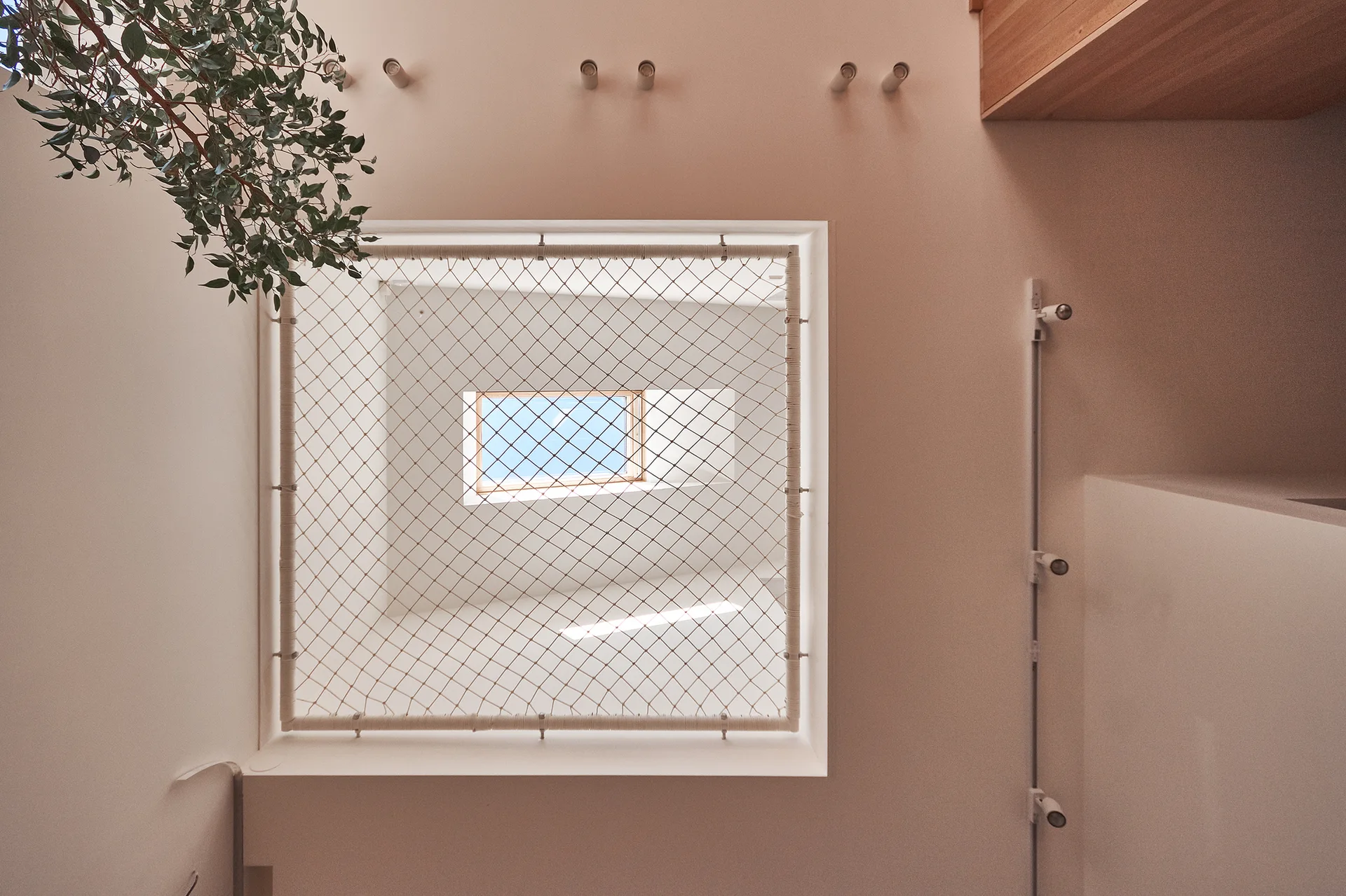 A low-angle view of a white rope safety net secured over a floor opening beneath a skylight in a minimalist beige interior.
