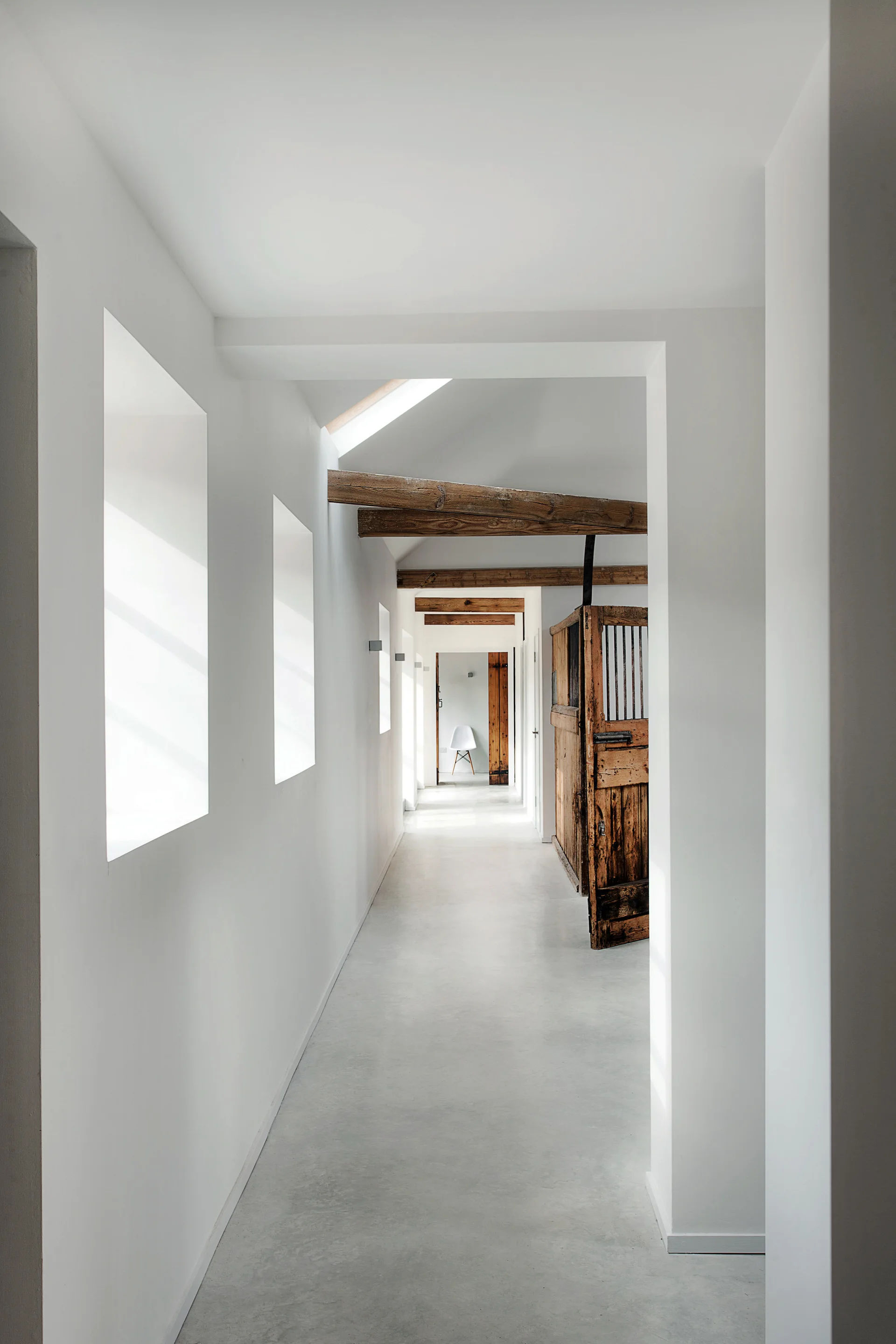 A long white minimalist hallway features exposed wooden ceiling beams, a rustic weathered wood stable door, and grey concrete flooring.