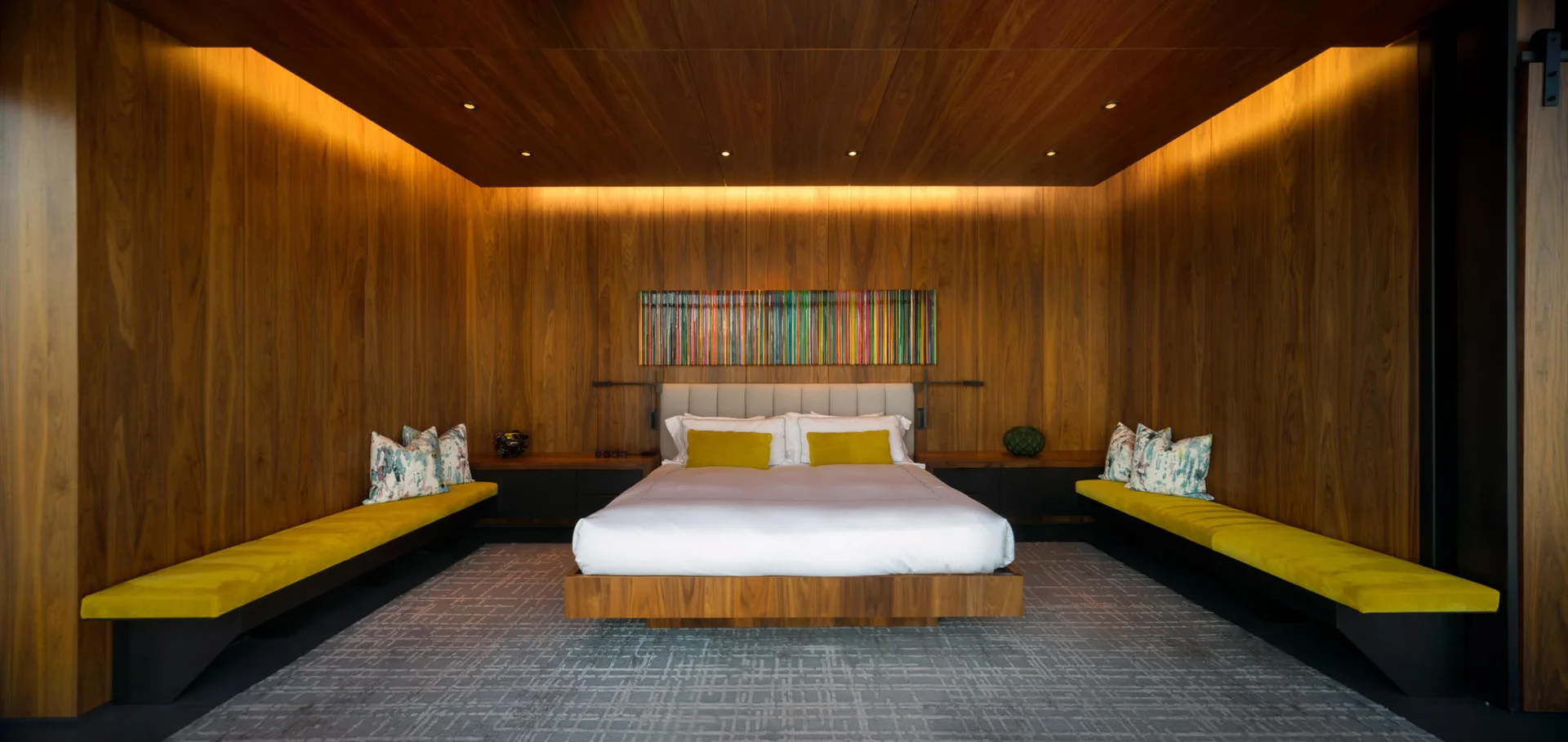 A contemporary bedroom with warm wood paneling on the walls and ceiling, a low bed, and long upholstered yellow benches.
