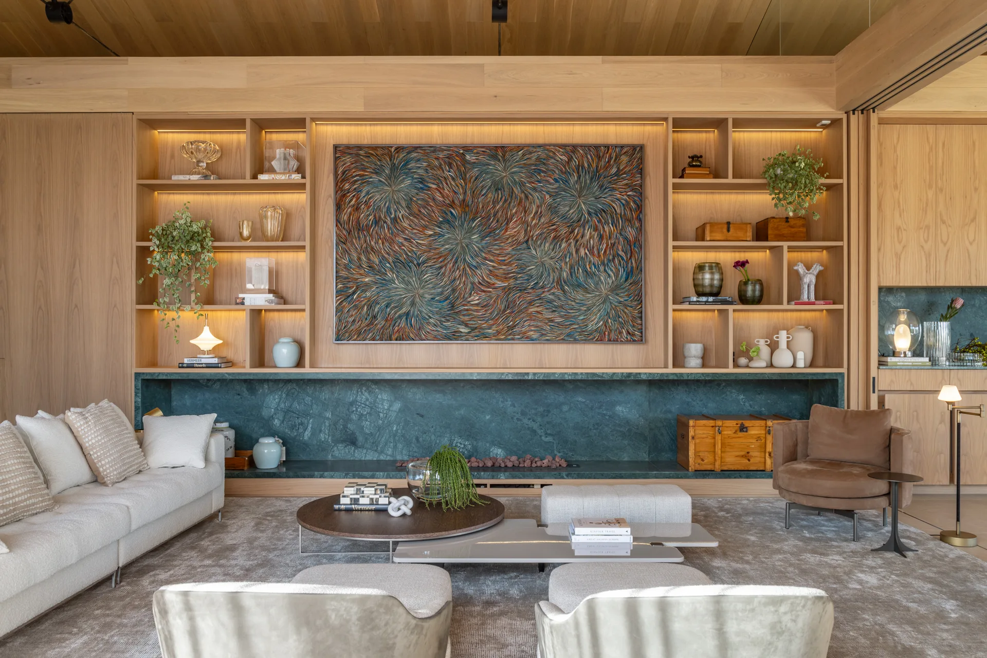 A modern living room features a warm wood paneled wall with built-in shelving, a green marble fireplace, a cream sofa, and low coffee tables.