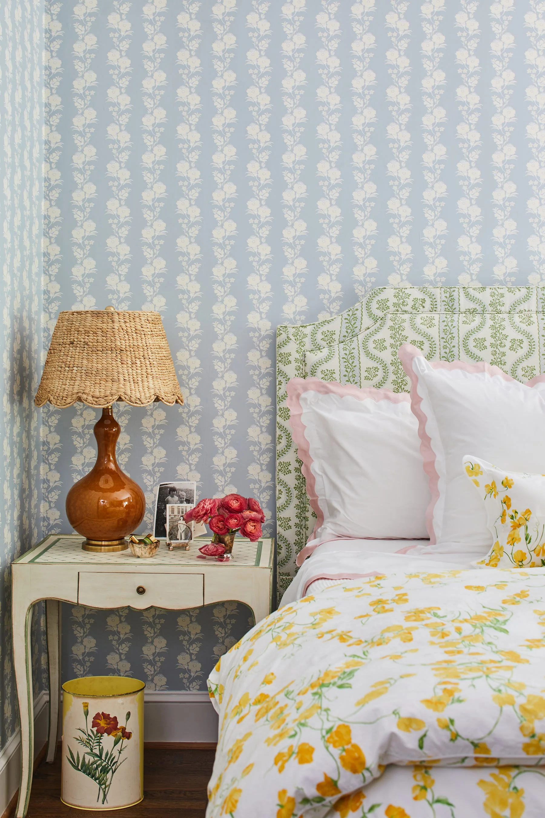 A cozy bedroom corner features blue floral wallpaper, an upholstered green headboard, and a bedside table with a scalloped woven lampshade.