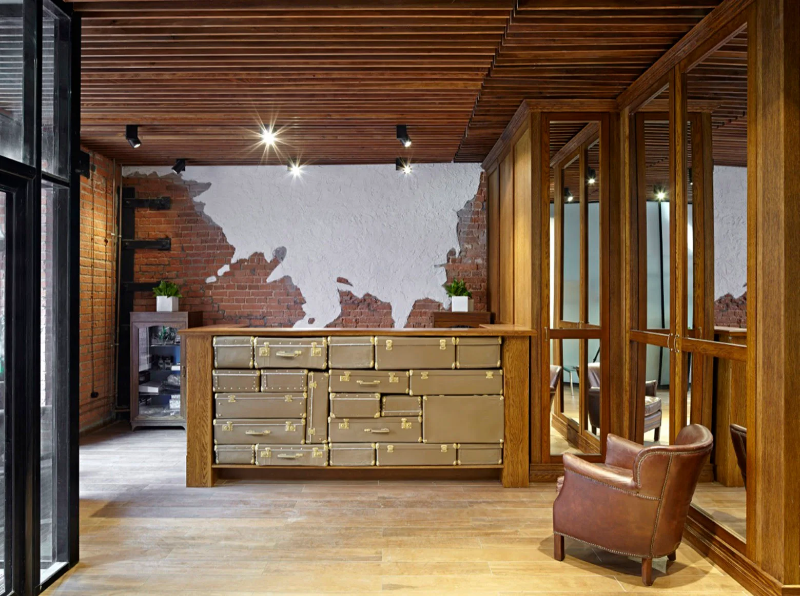 An industrial lobby features a wooden reception desk with suitcase-style drawers, set against an exposed red brick wall and a wood slatted ceiling.