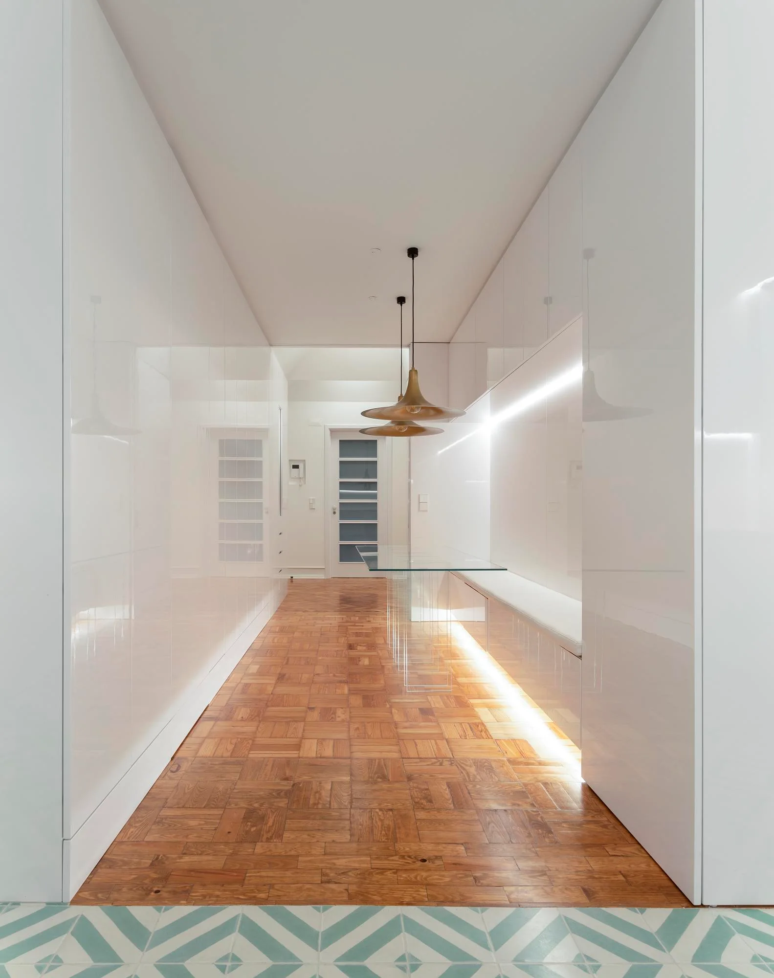 A minimalist white hallway featuring high-gloss built-in cabinetry, warm wood parquet flooring, integrated LED lighting, and two brass pendant lights.