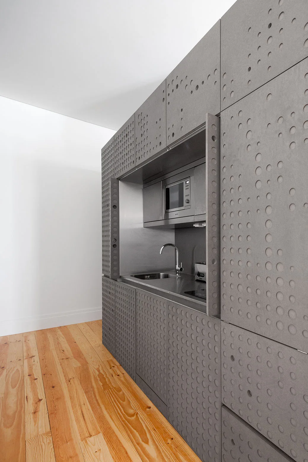 A compact kitchen nook featuring stainless steel appliances and a sink is recessed within grey cabinets with a perforated circular pattern.
