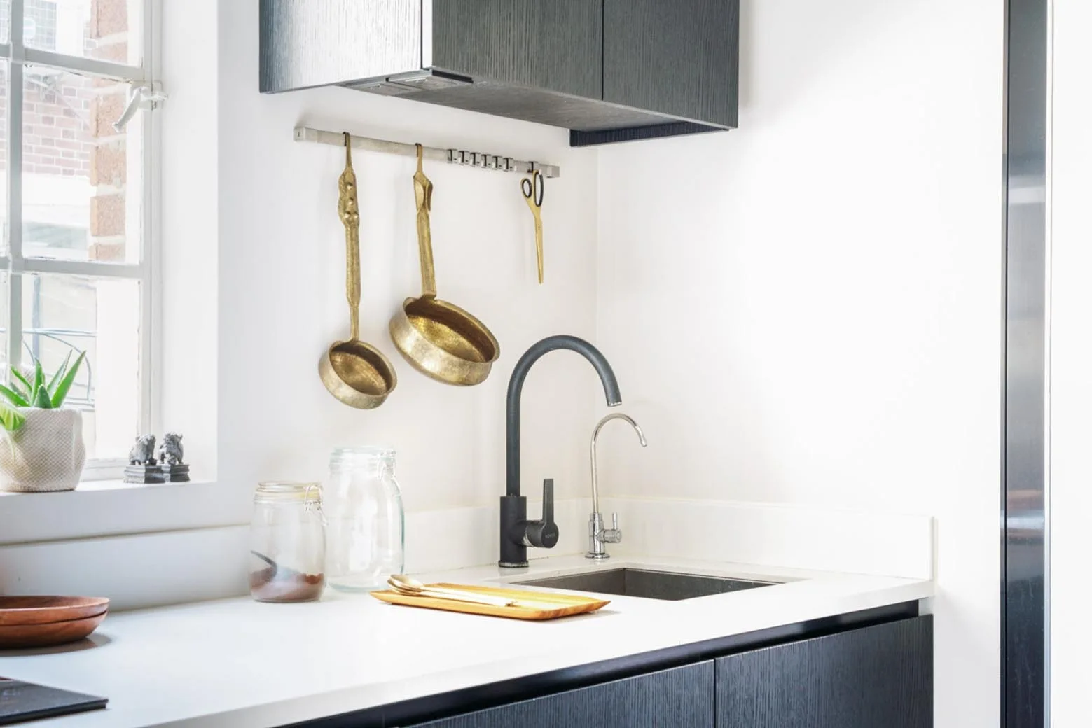 A modern kitchen corner features a black faucet, white countertops, dark cabinetry, and brass utensils hanging on a silver wall rail.