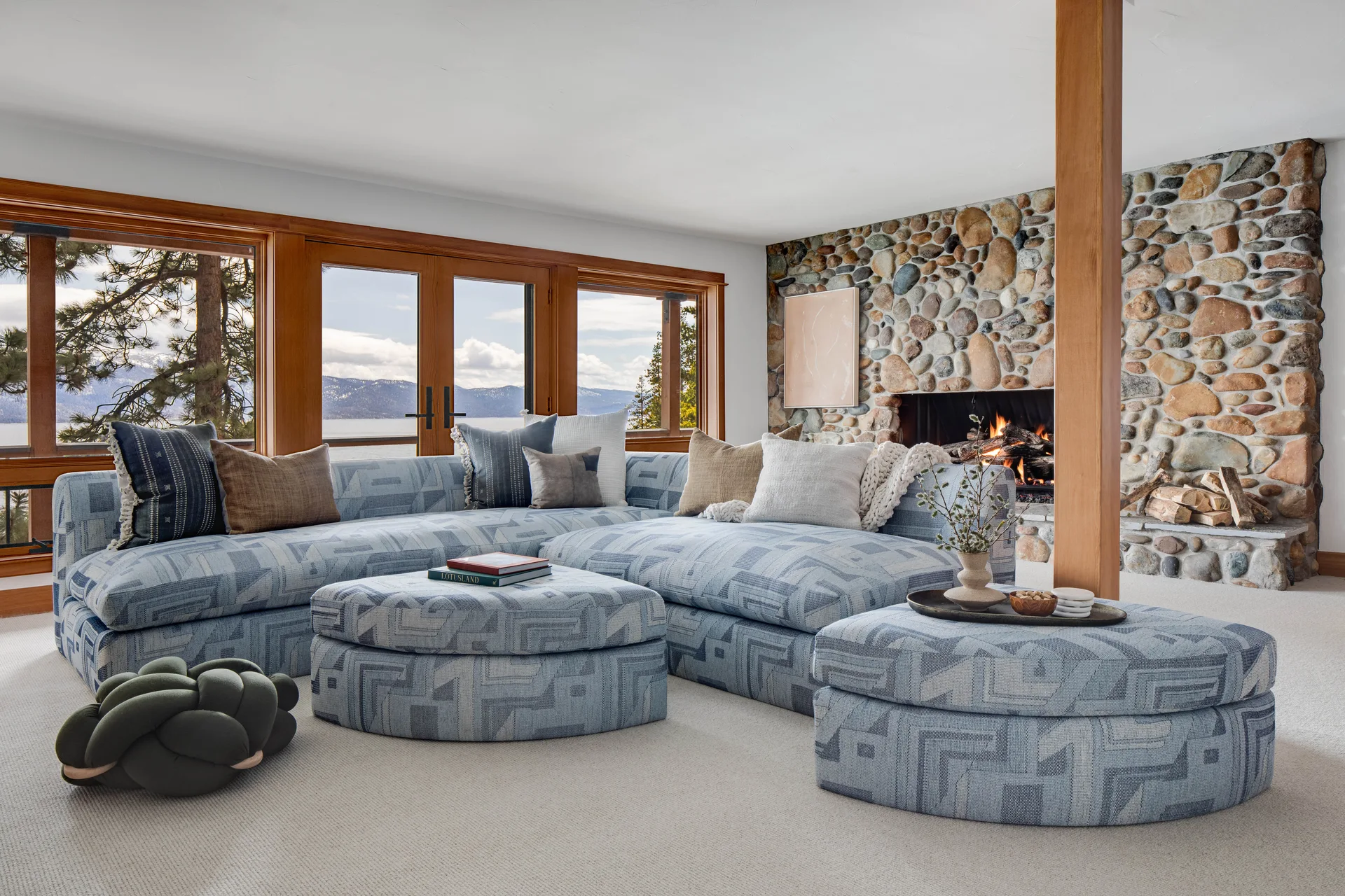 A living room featuring a blue patterned sectional sofa, matching round ottomans, a stone fireplace, and wood-framed windows looking out to mountains.