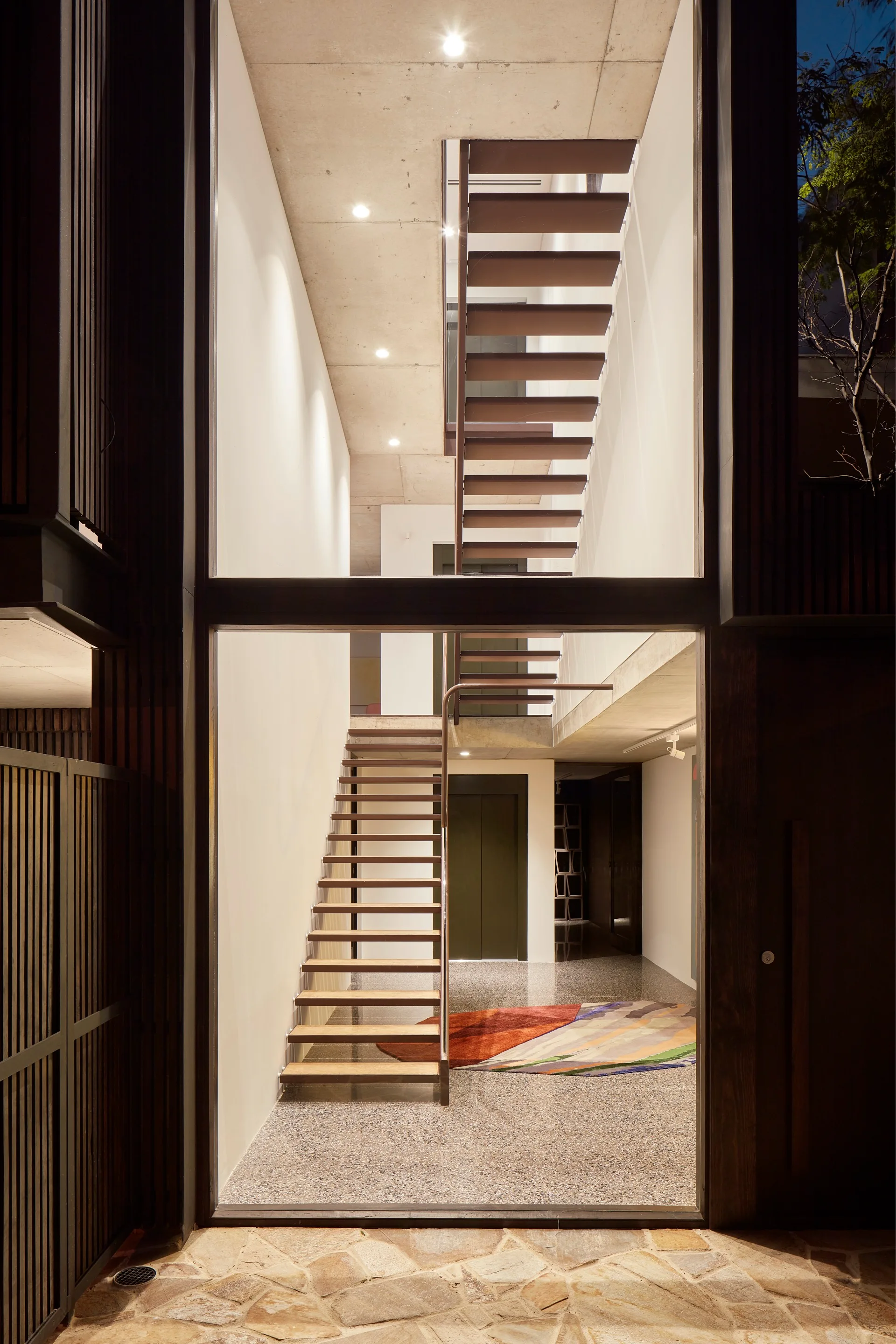 A modern multi-story entryway viewed through glass, featuring a steel open-riser staircase, white walls, and a grey terrazzo floor.