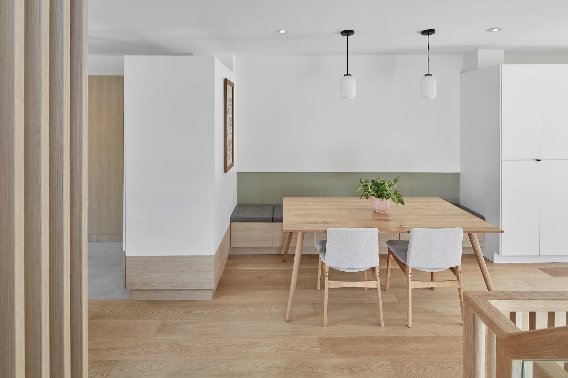 A light wood dining table with gray chairs and a built-in corner banquette sits before a white wall with a painted sage green stripe.