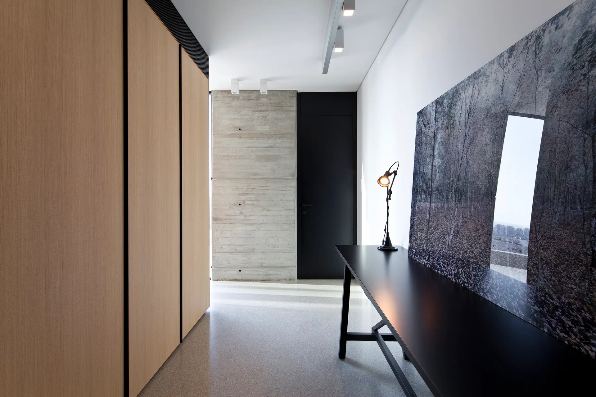 A minimalist hallway with full-height light wood cabinets, a raw concrete wall, and a black console table with a leaning art piece.