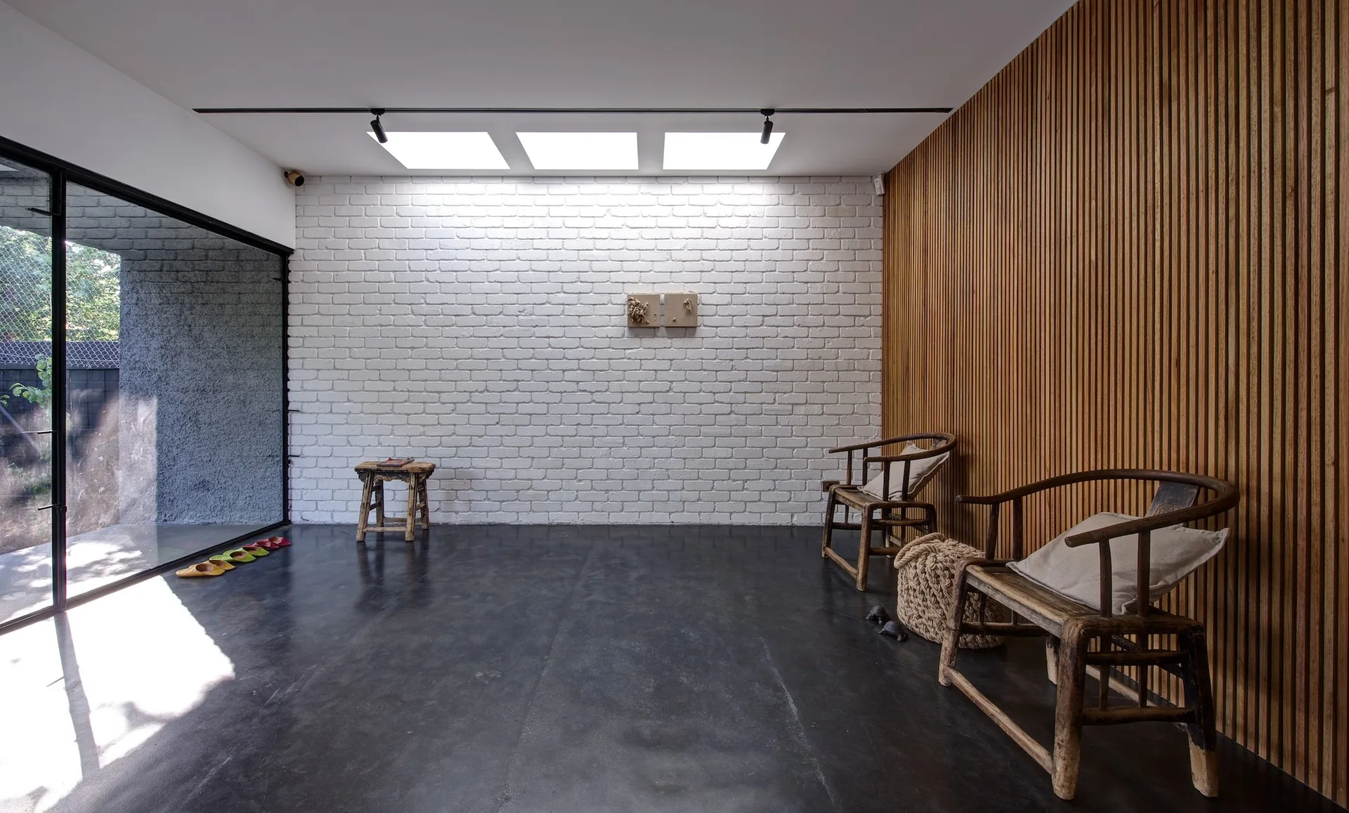 A minimalist room features a white painted brick wall, a vertical wood slatted wall, and two rustic wooden chairs lit by three skylights.