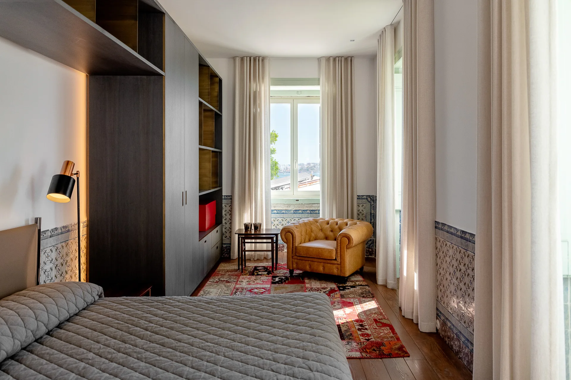 A bedroom featuring a grey quilted bed, a large dark wardrobe, a tan tufted leather armchair by a window, and patterned blue and white wall tiles.