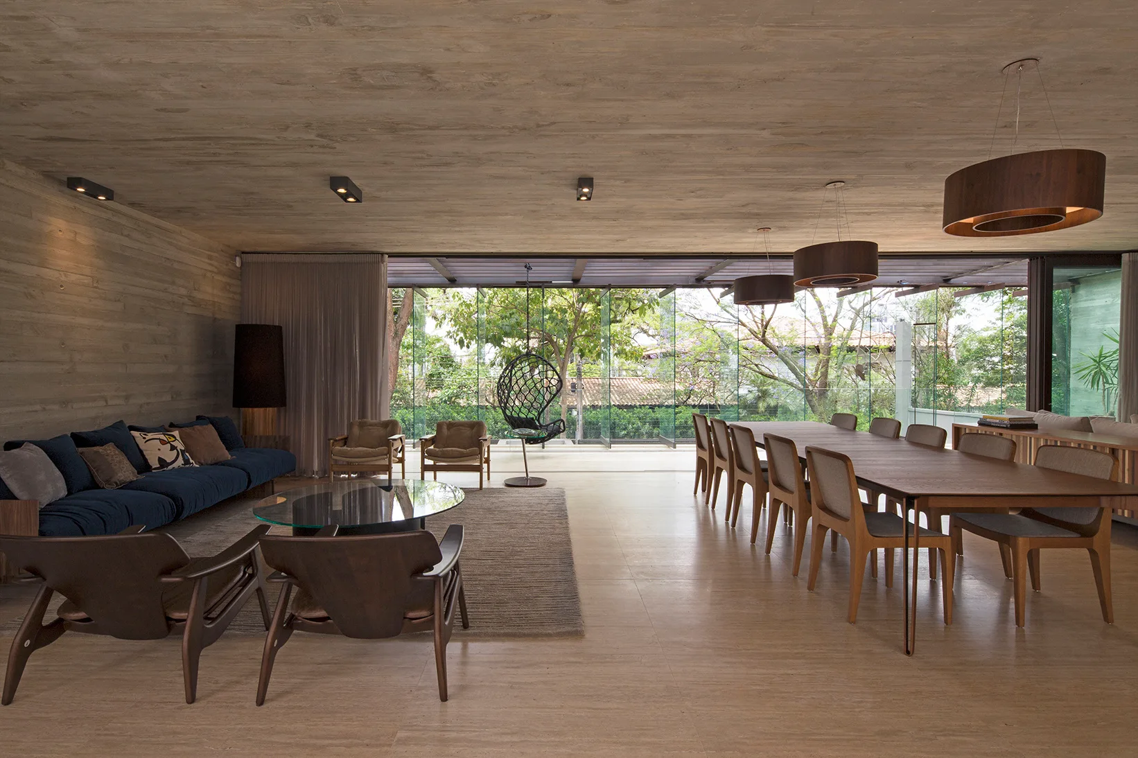 An open-plan living and dining space with a long wooden table, a blue sofa, and board-formed concrete ceilings.