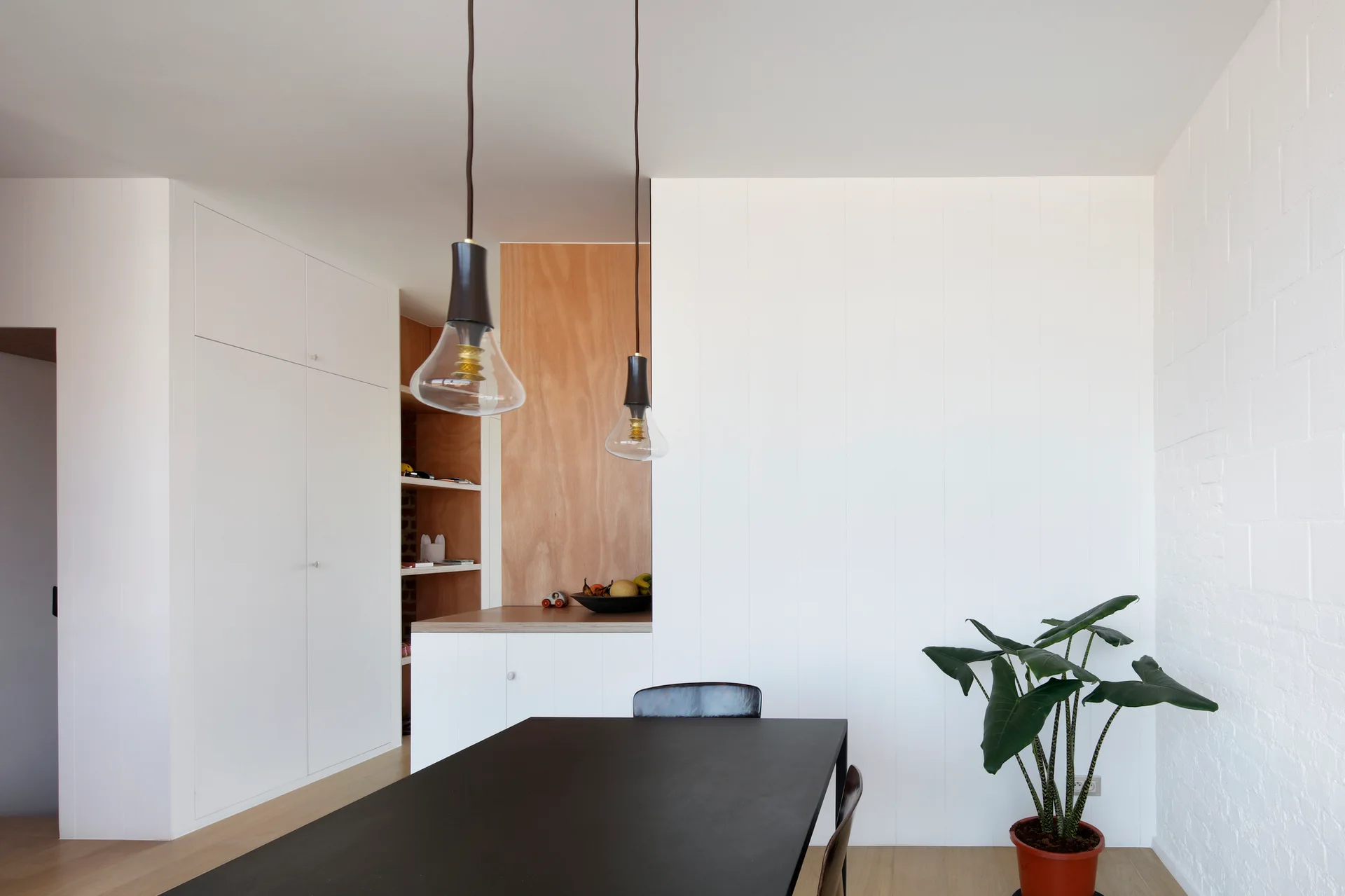 A minimalist dining room features a black dining table, glass pendant lights, white paneled walls, and a recessed natural wood shelving niche.