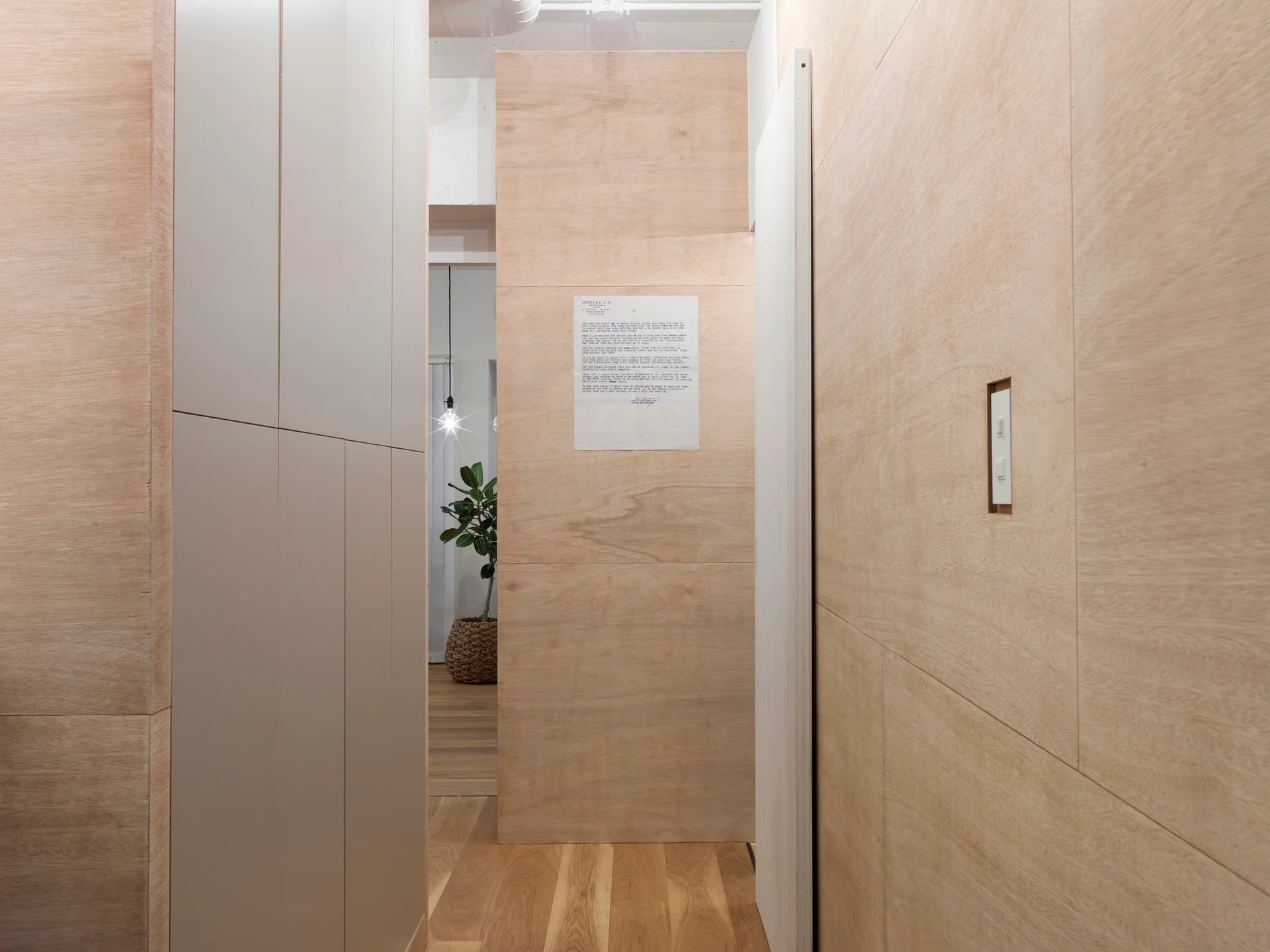 A minimalist hallway features light wood veneer wall panels, a tall off-white cabinet, and a doorway looking into another room.