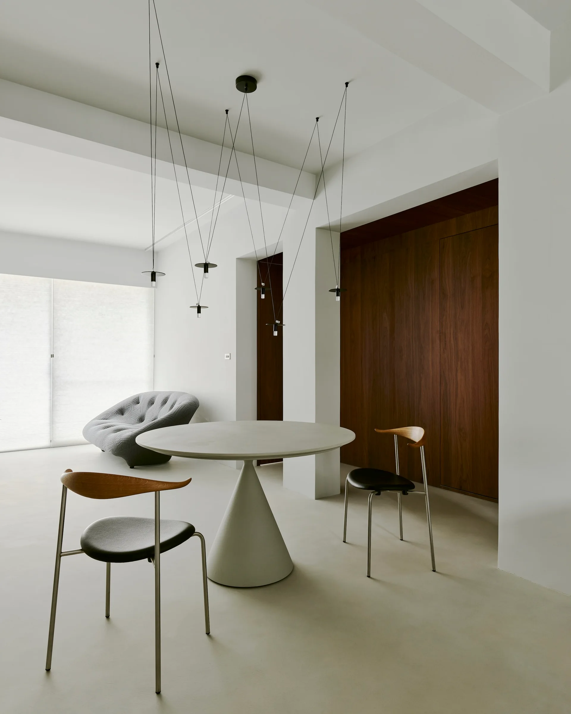 A minimalist dining room features a round gray pedestal table, two wood-accented chairs, and a black multi-cable pendant light fixture.