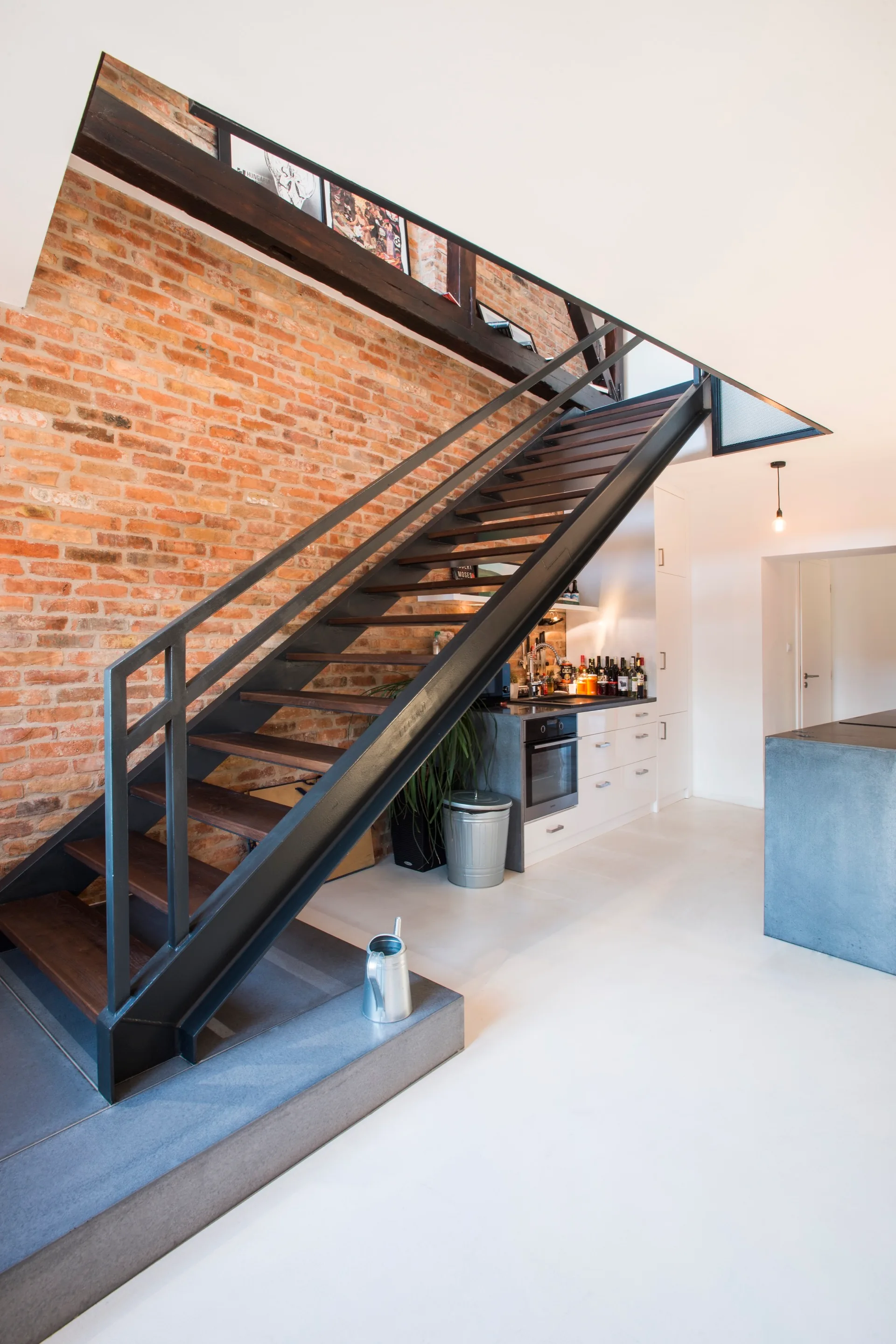 An industrial black metal staircase with wood treads stands next to an exposed red brick wall in a loft space with a minimalist kitchen.