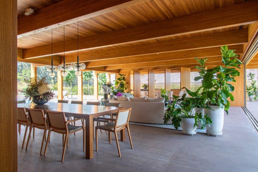 An open-concept dining area features a large wood table, mid-century chairs, a wood-paneled ceiling with beams, and glass doors opening to the outdoors.