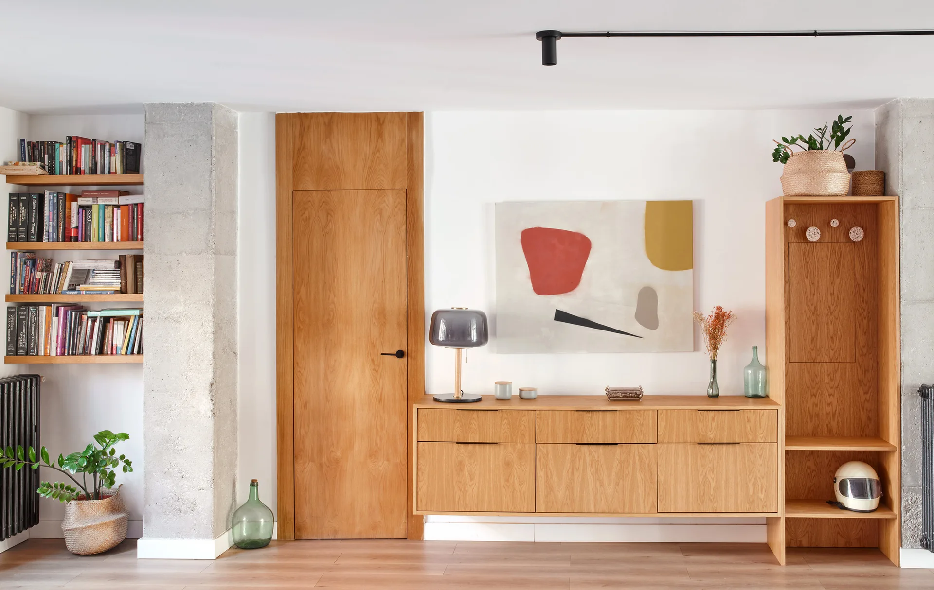 A wooden sideboard, matching flush door, and tall shelving unit stand against a white wall flanked by exposed concrete pillars.