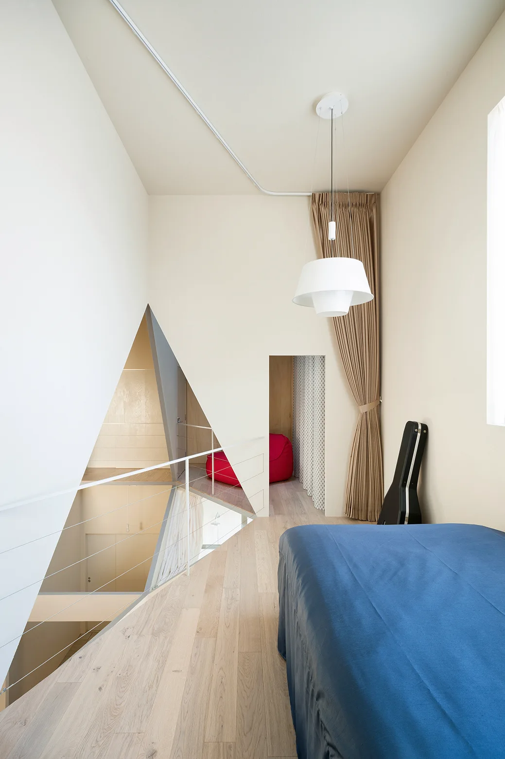 A minimalist loft bedroom features light wood flooring, a blue bed, and a large triangular cutout overlooking the level below.