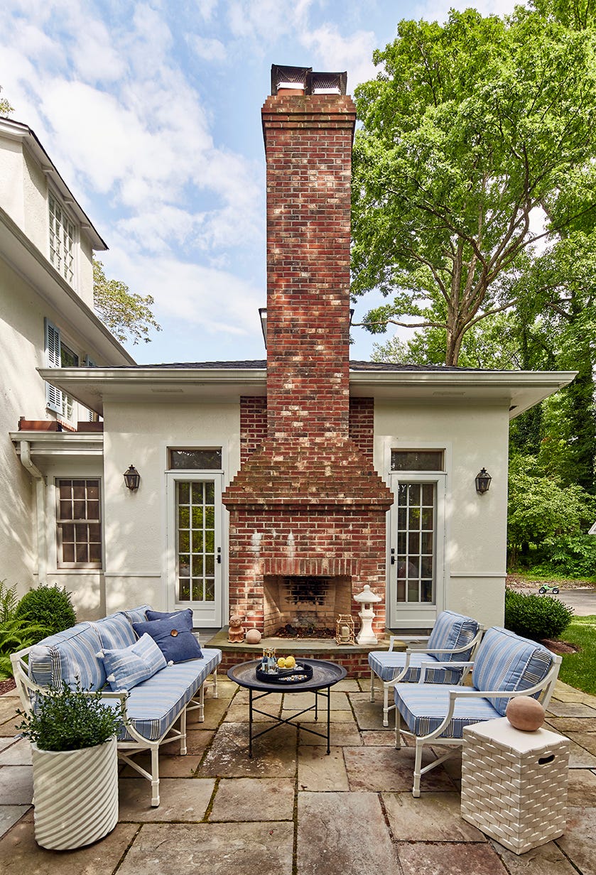 A stone patio features a tall red brick fireplace flanked by white glass doors and comfortable blue and white striped outdoor furniture.