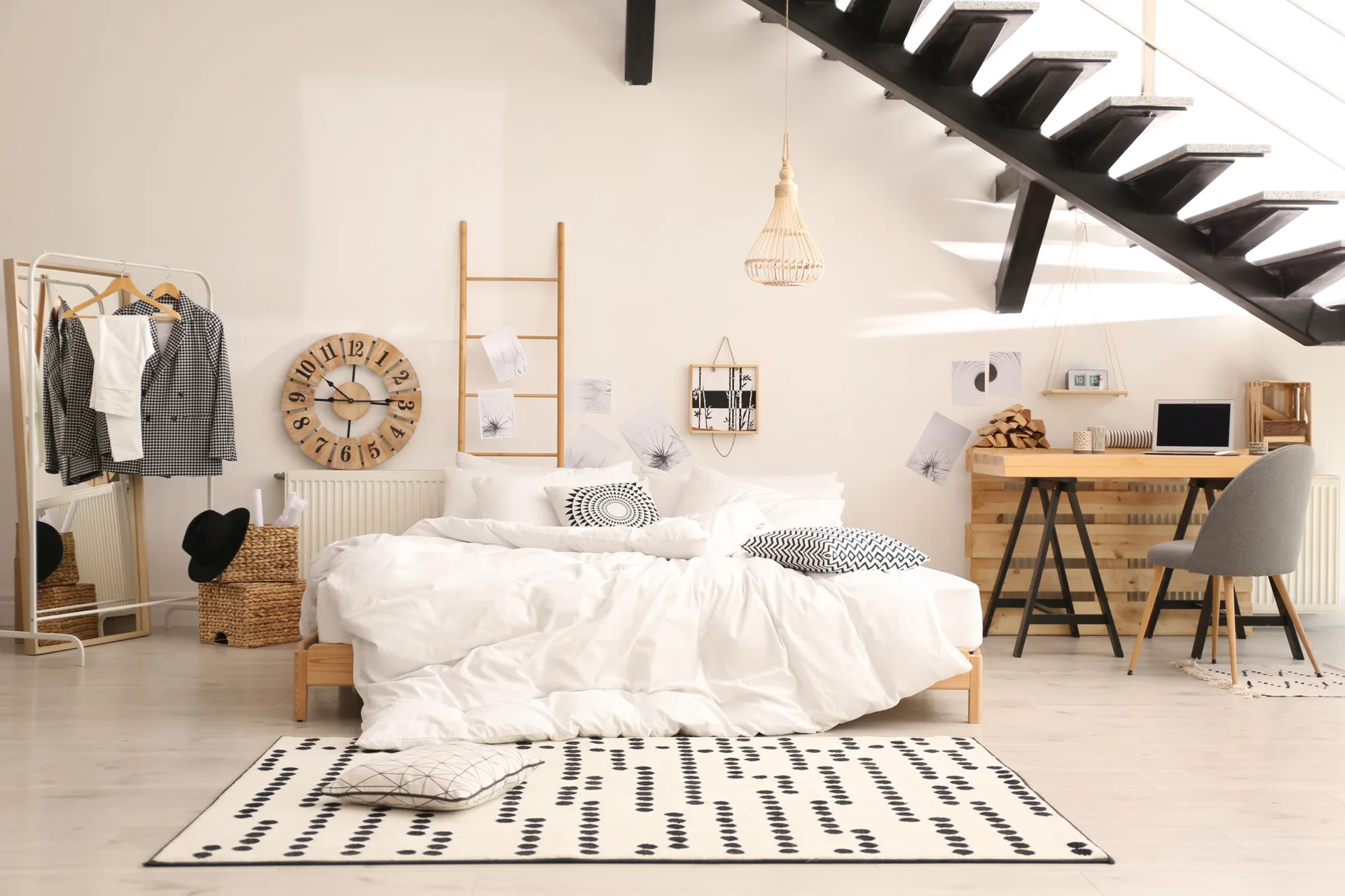 A minimalist bedroom features a low wooden bed with white linens, a wooden desk, and a black open staircase against a white wall.