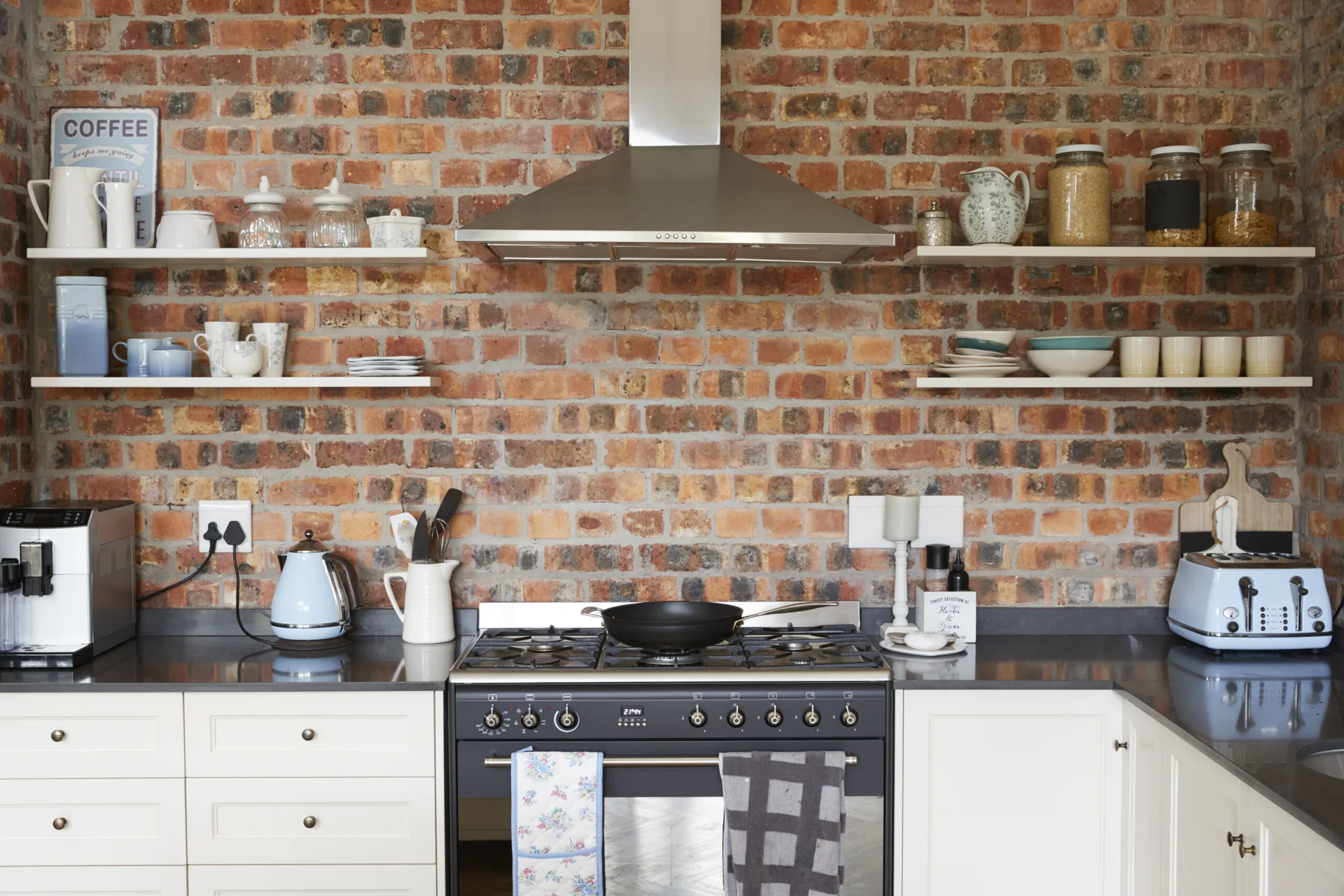 A rustic kitchen features a red brick wall, white floating shelves, a stainless steel range hood, and light blue appliances on a dark countertop.