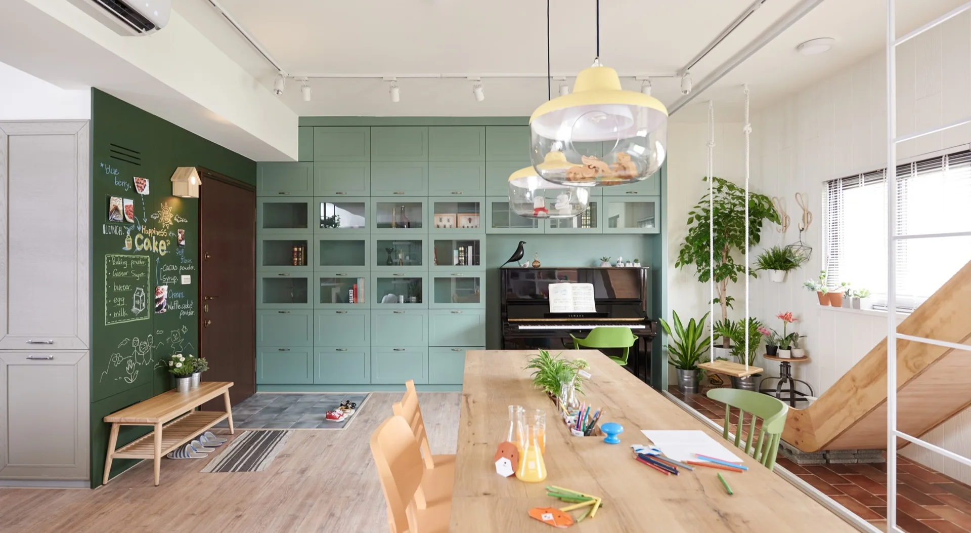 A bright family living area features a large sage green storage wall, a wooden dining table, a piano, and an indoor wooden slide.