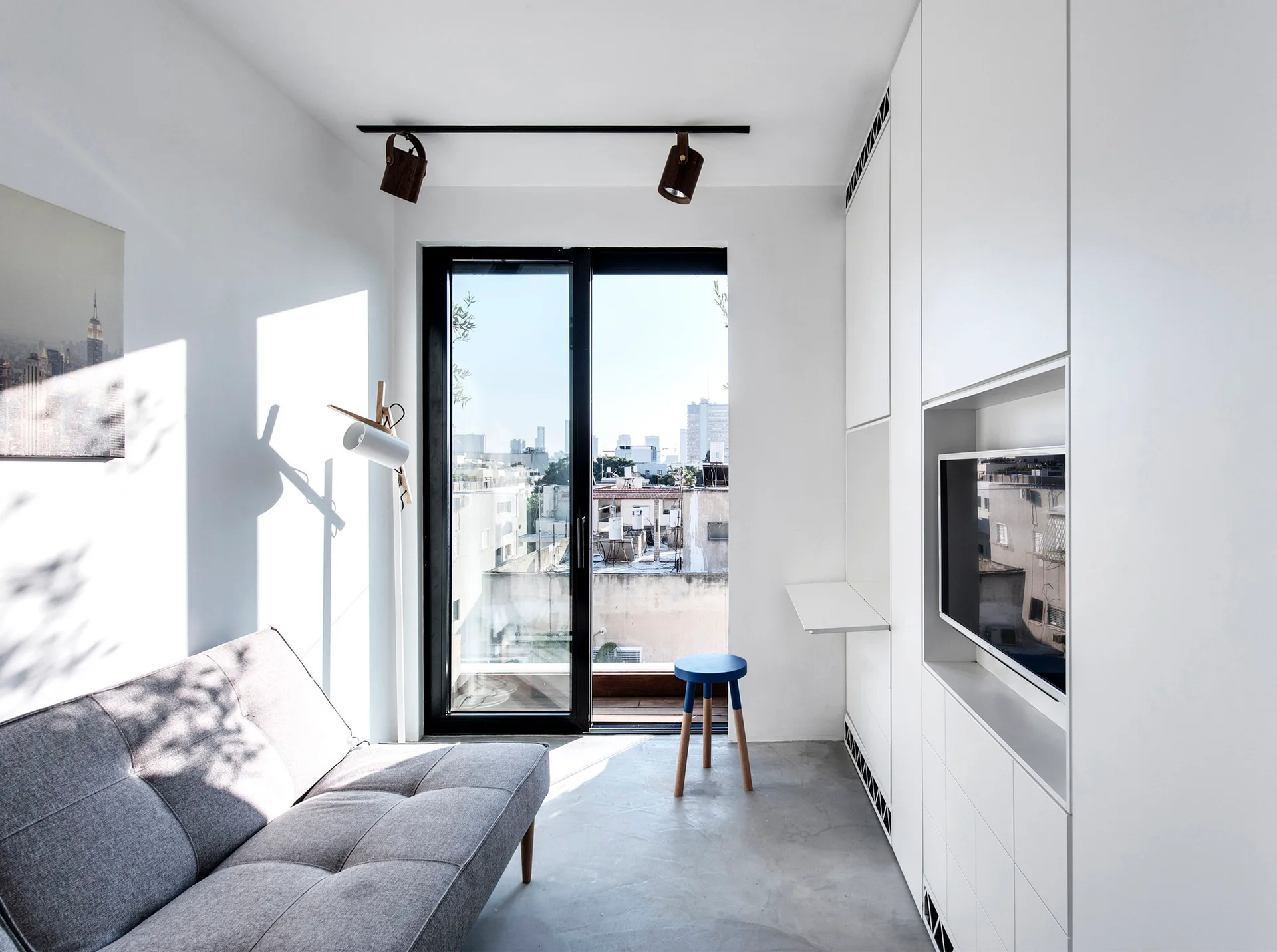 A minimalist living room featuring a grey sofa, a polished concrete floor, and a full-height white storage wall with an integrated television.