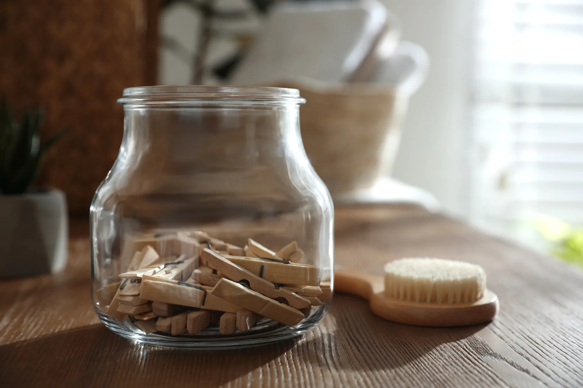A clear glass jar filled with wooden clothespins rests on a warm wood tabletop next to a wooden cleaning brush in natural light.
