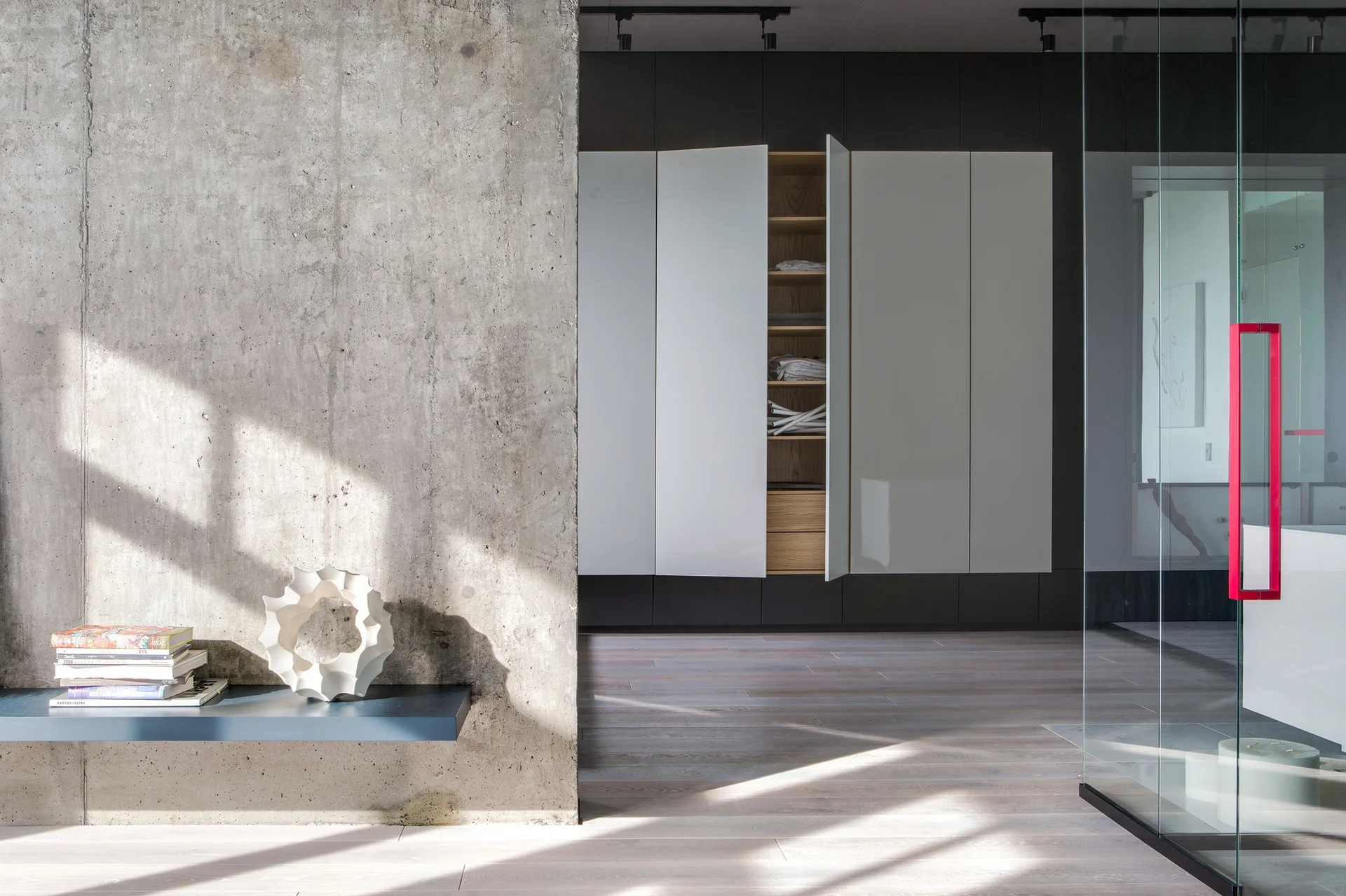 A modern room features a textured concrete wall with a floating shelf, a glossy white built-in wardrobe, and a glass partition with a red handle.