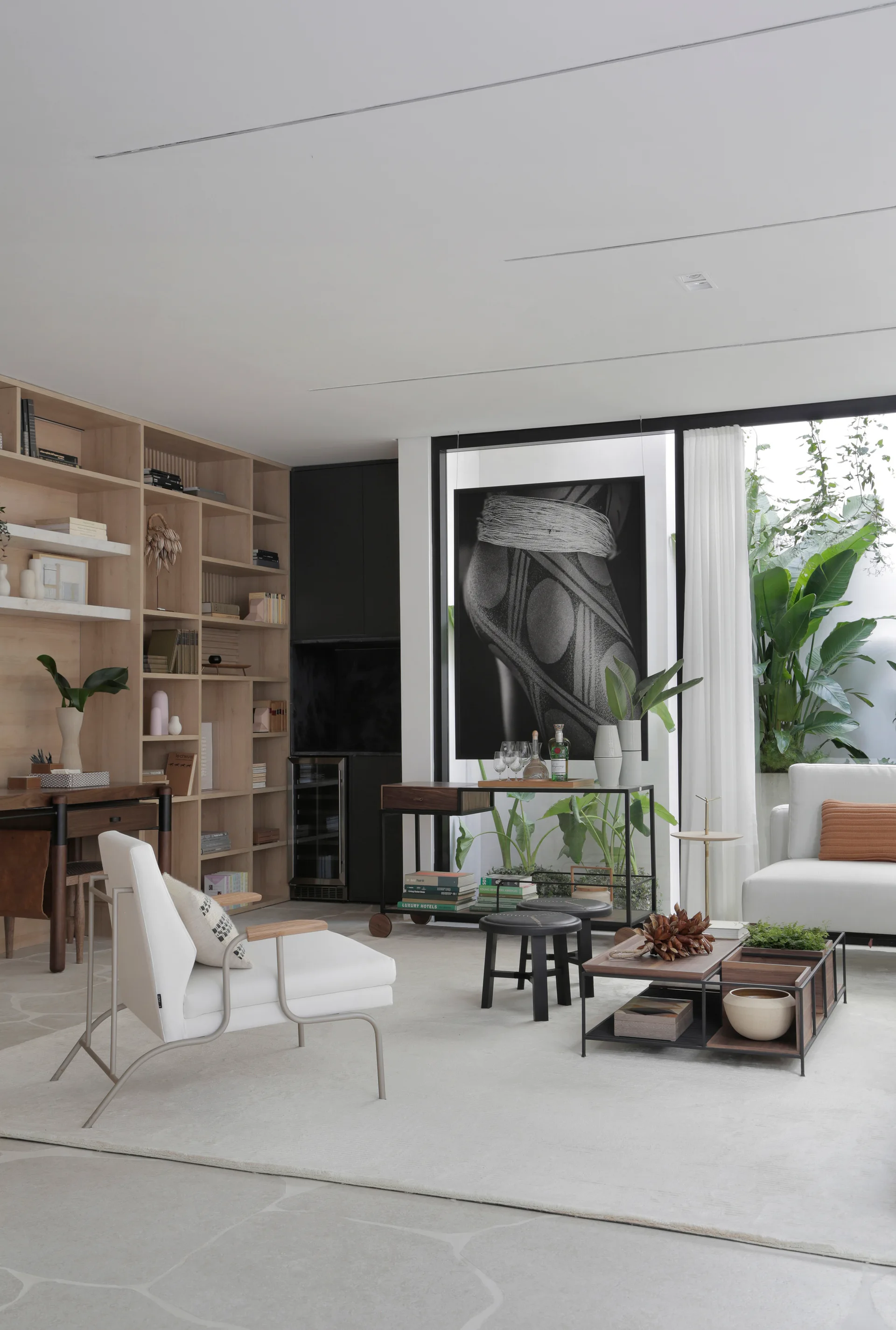 A bright living room features a large light oak built-in shelving unit, a white contemporary armchair, a modular coffee table, and large glass windows.