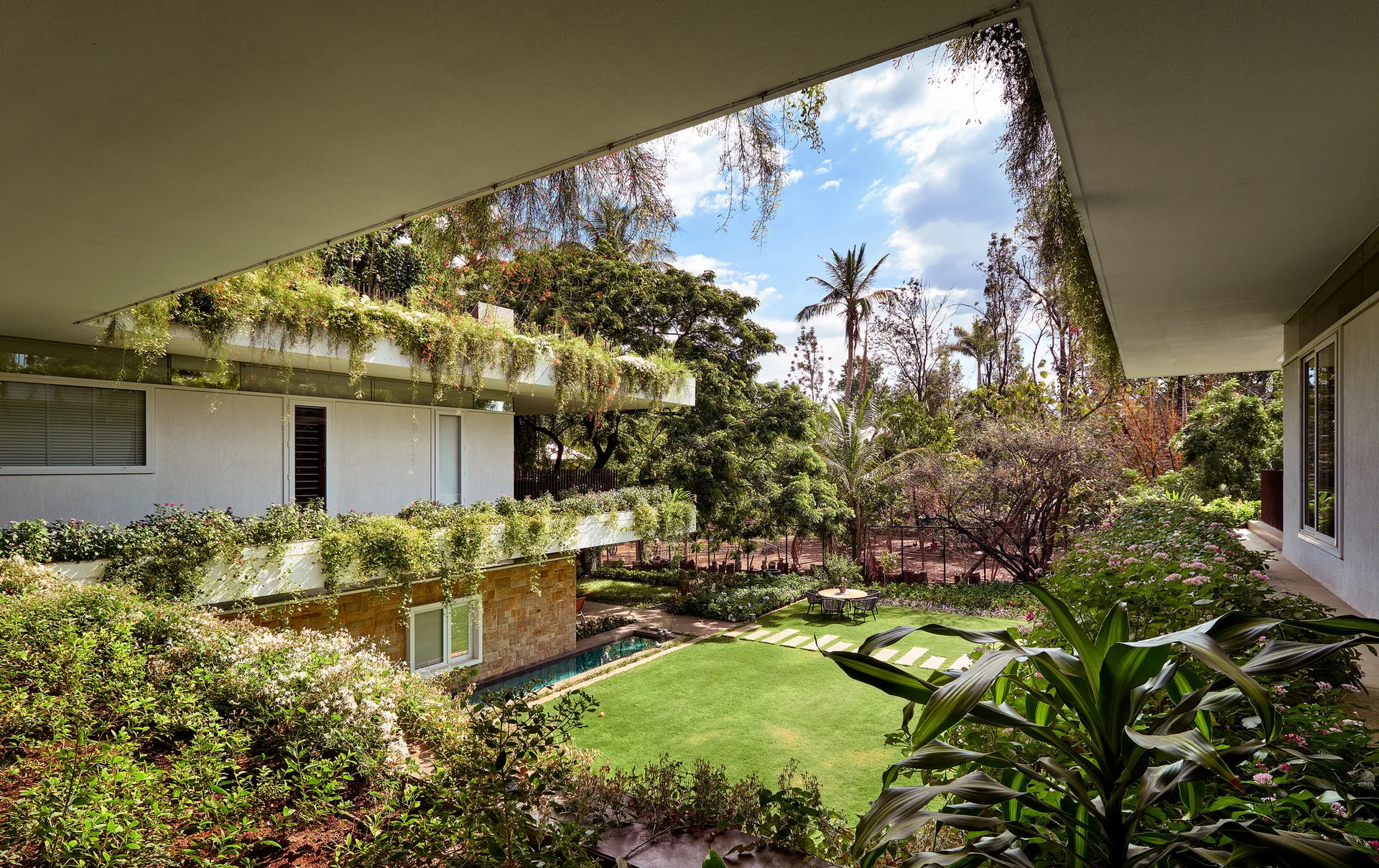 A framed view from a shaded patio looking out at a modern house with white walls, stone cladding, and balconies spilling with lush green plants.