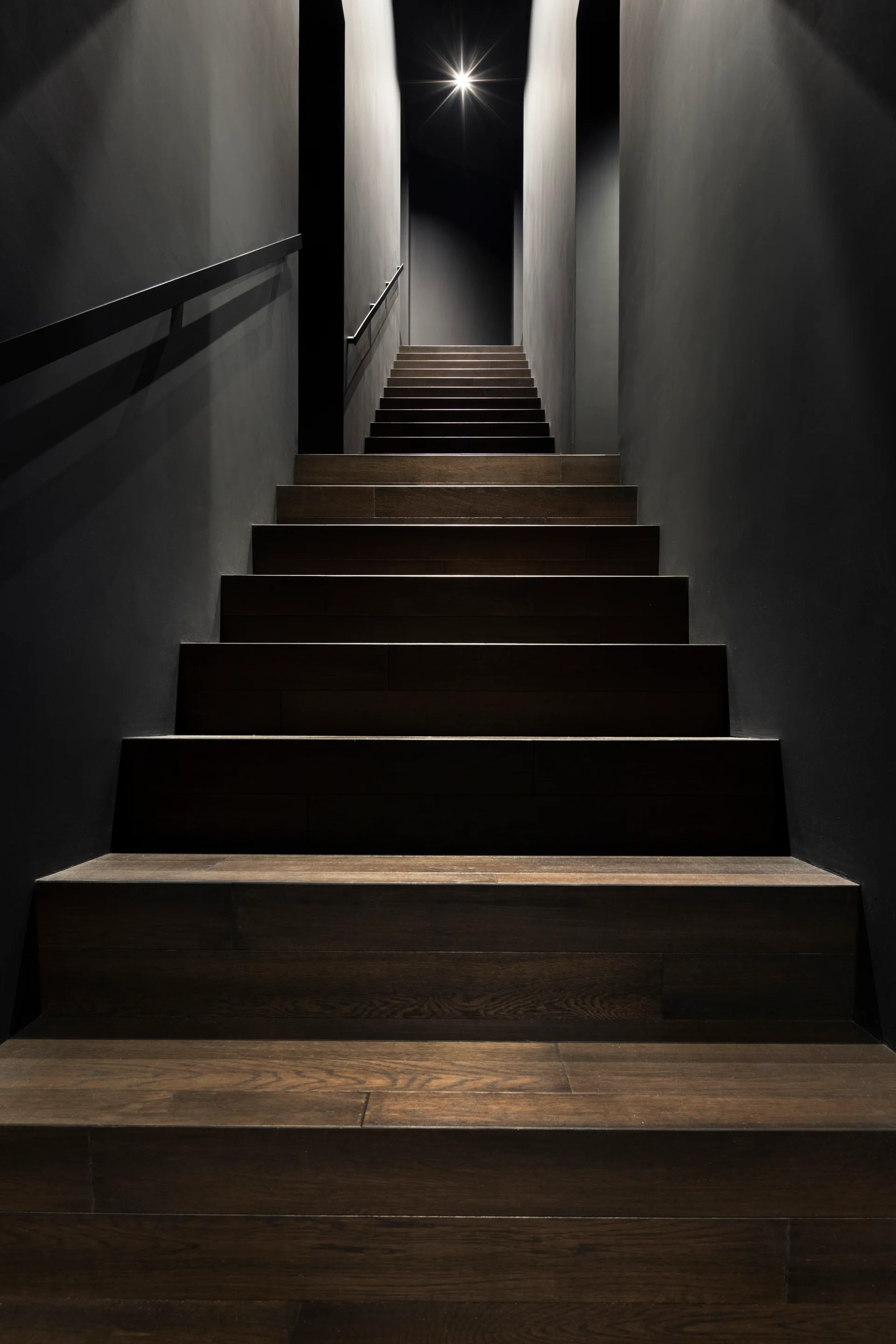 A modern staircase features dark wood steps, matte charcoal walls, a sleek black handrail, and a single bright spotlight shining from the upper landing.
