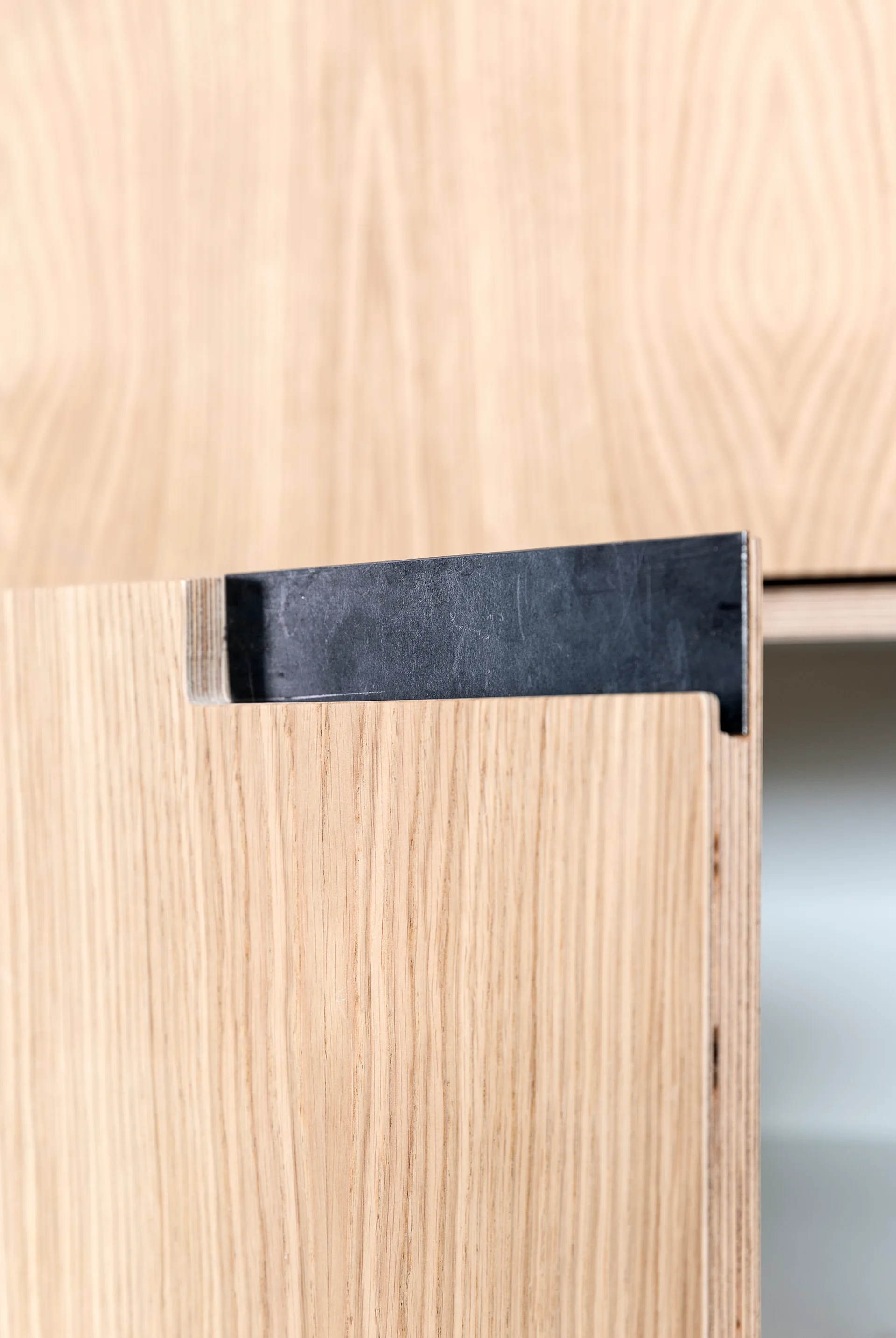 A close-up of a light oak plywood cabinet door showing an integrated black metal pull handle and exposed layered edge.
