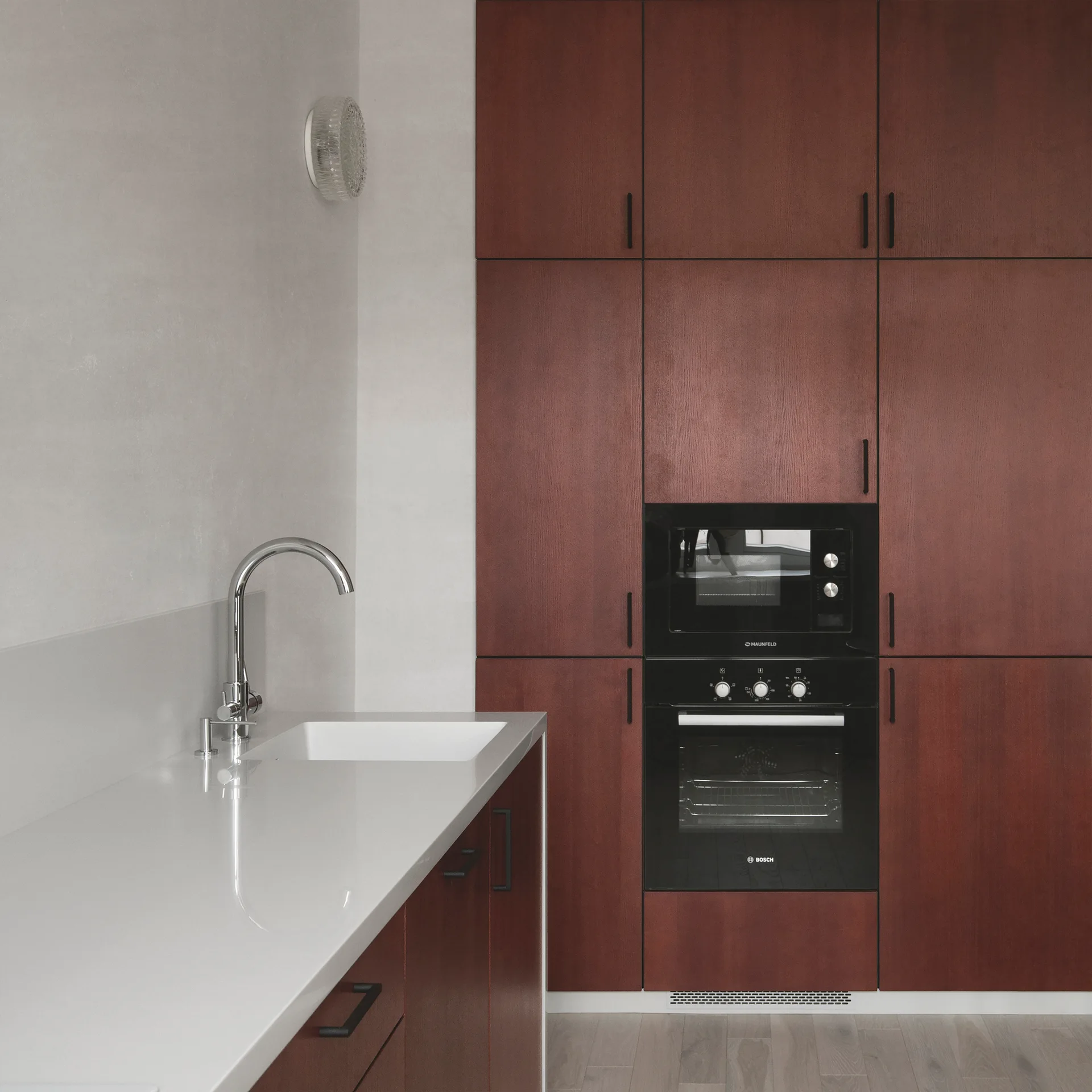 A modern kitchen featuring deep red-brown wood cabinets, integrated black appliances, a light grey countertop, and a chrome gooseneck faucet.