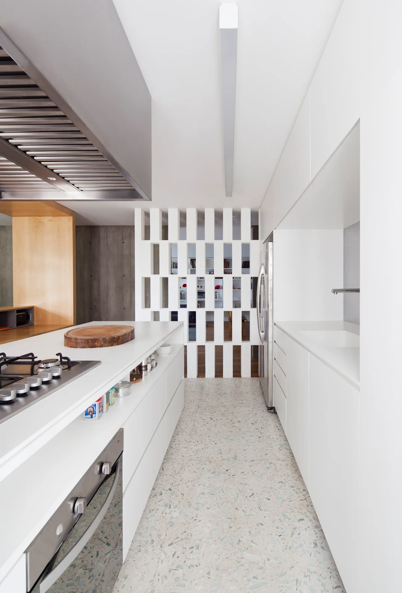 A minimalist white galley kitchen featuring a central island with a cooktop, speckled terrazzo flooring, and a white slatted partition wall.