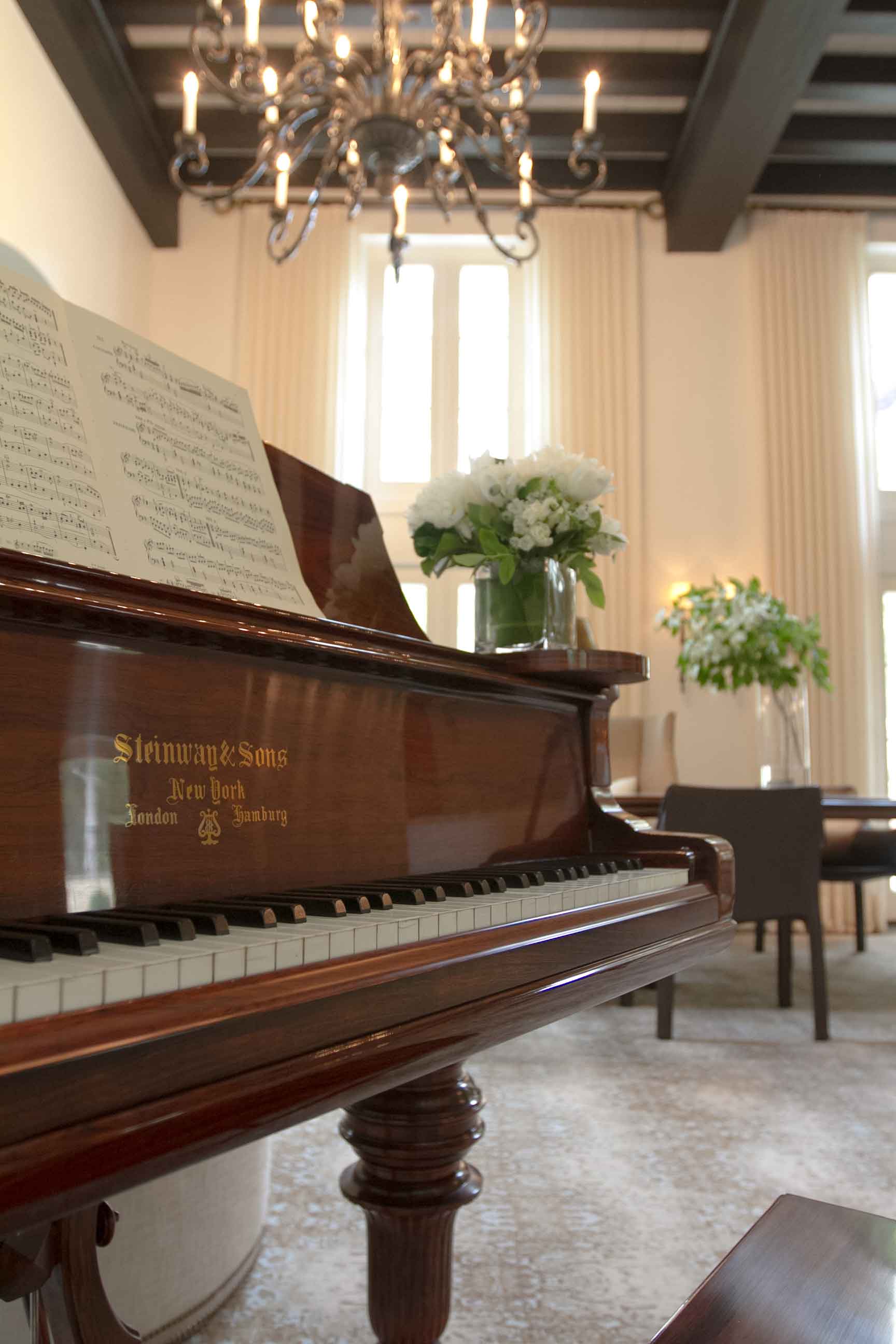 A close-up of a polished dark wood grand piano with sheet music and white flowers, set in a room with exposed ceiling beams and a chandelier.