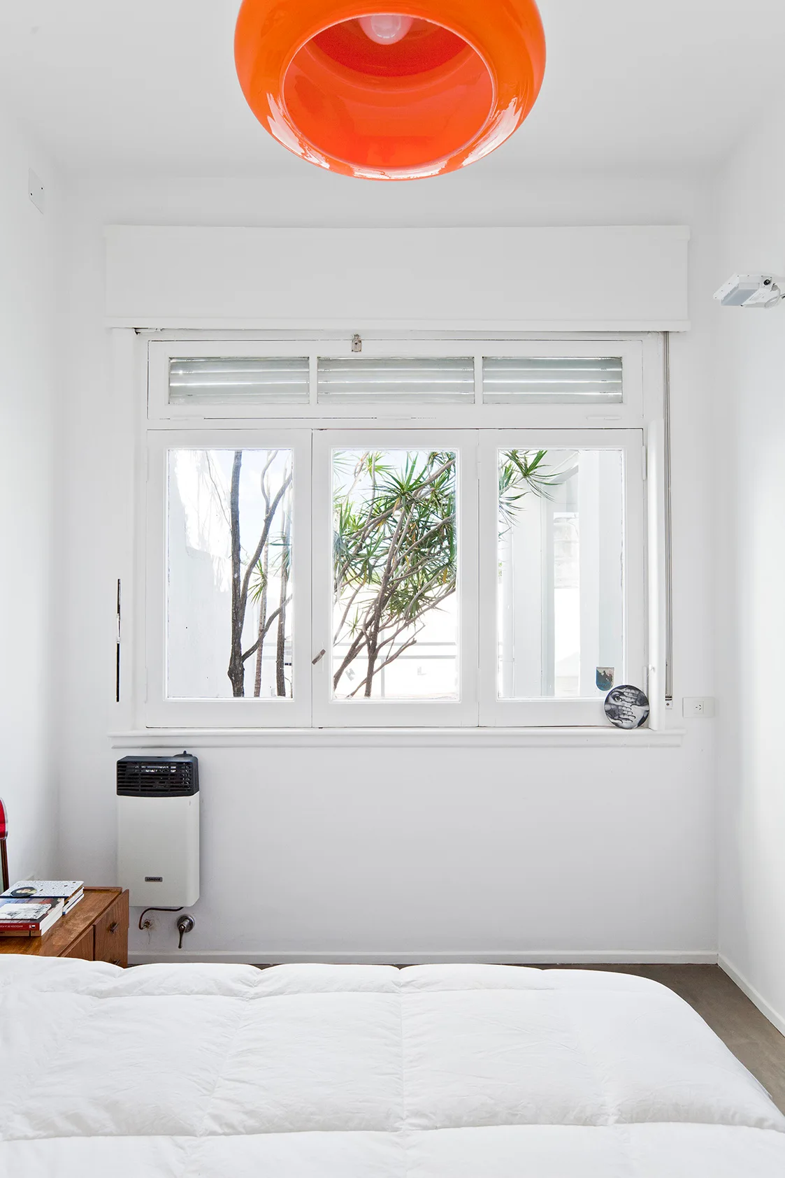 A minimalist white bedroom features a puffy white duvet on the bed, a large white window showing green plants, and a bright orange ceiling light.