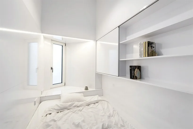 A small, minimalist white bedroom featuring a built-in bed, recessed strip lighting, and a wall of integrated white shelving.