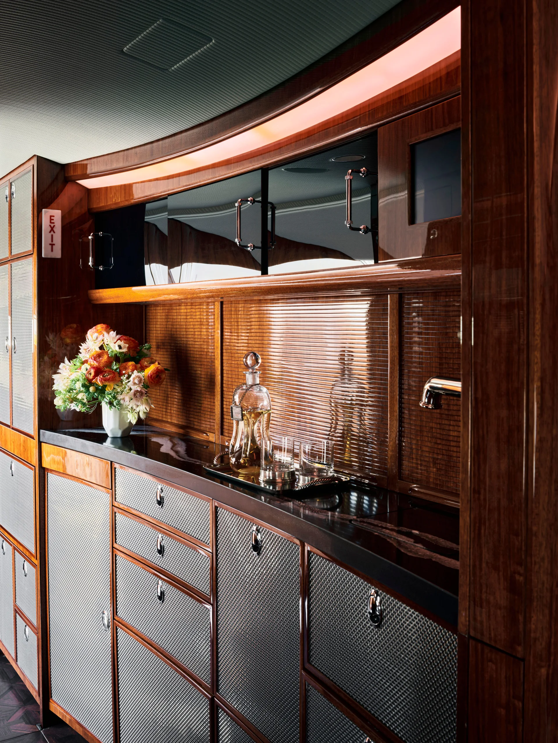 A polished wood bar cabinet with woven silver metal mesh door panels, integrated ambient lighting, and glassware on a counter.