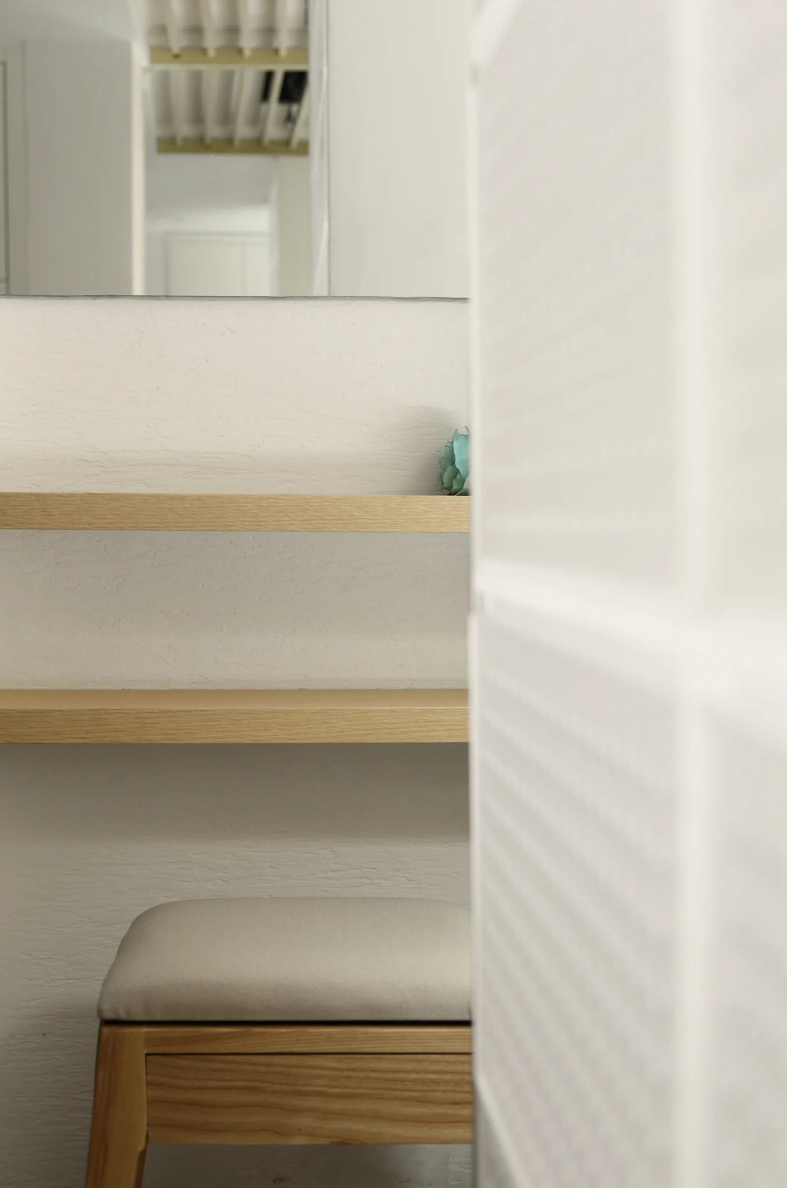 A minimalist dressing nook features two floating light oak shelves mounted on a textured plaster wall above a padded wooden stool.